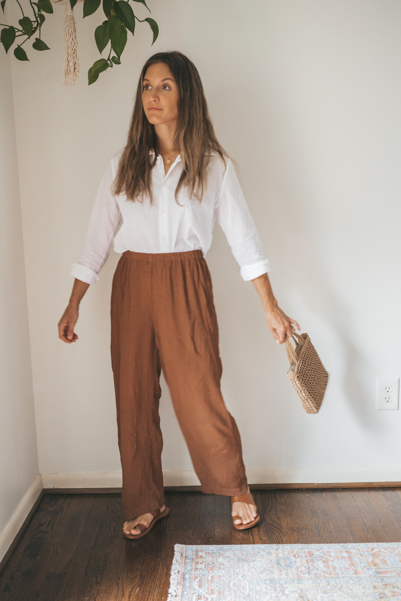 ashley wearing a white button down, brown drapey pants, brown leather sandals, and a woven bag