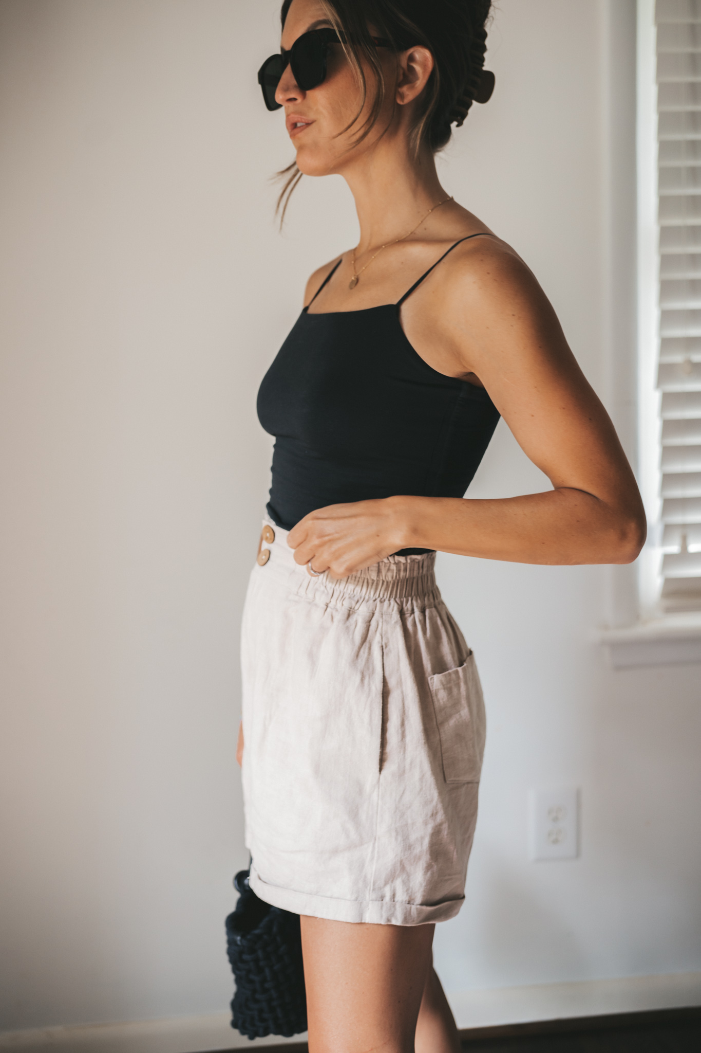 ashley wearing a black cropped tank, tan linen shorts, black strappy sandals, tortoise shell sunglasses and a black woven bag