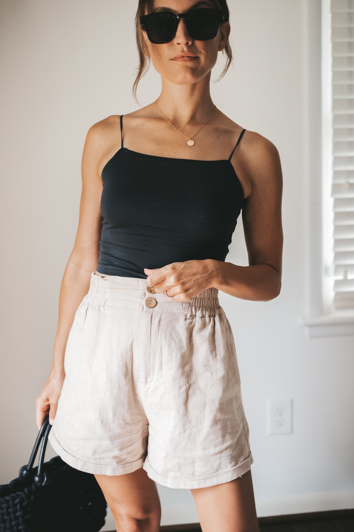 ashley wearing a black cropped tank, tan linen shorts, black strappy sandals, tortoise shell sunglasses and a black woven bag