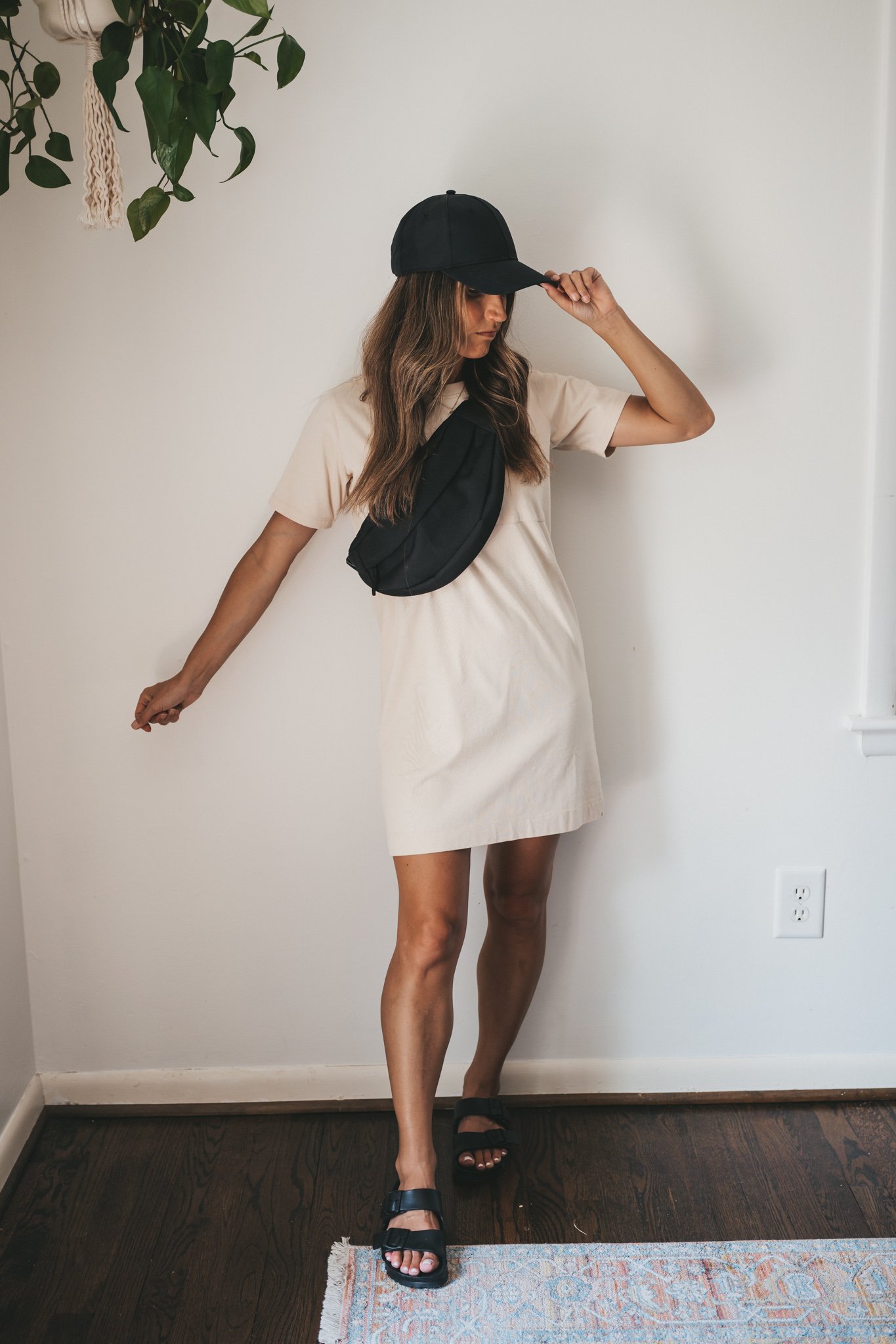 ashley wearing a cream colored t-shirt dress, black birkenstocks, a black belt bag, and a black baseball hat