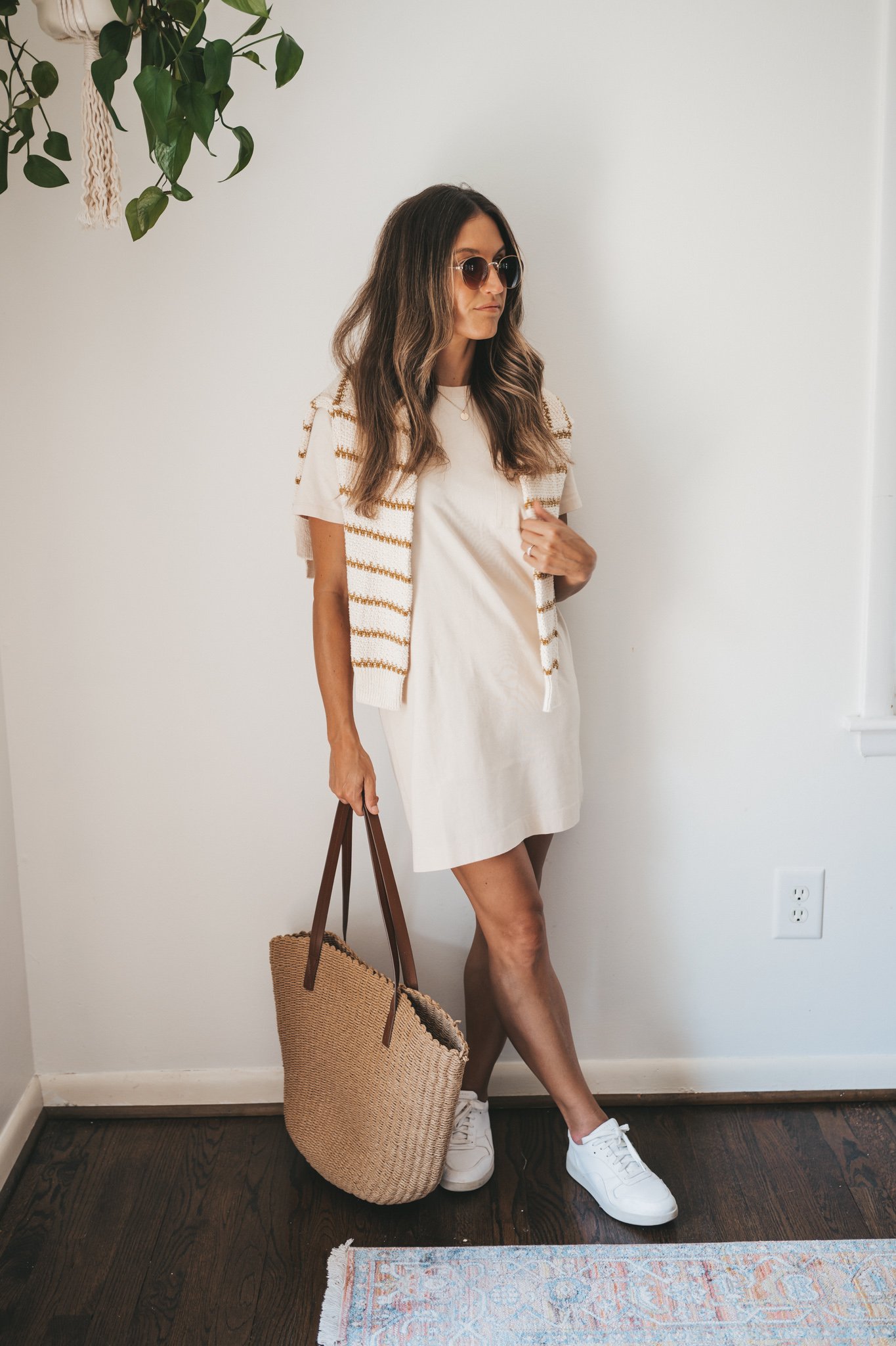 ashley wearing a cream colored t-shirt dress, striped sweater worn over the shoulders, white fashion sneakers, and a woven tote bag