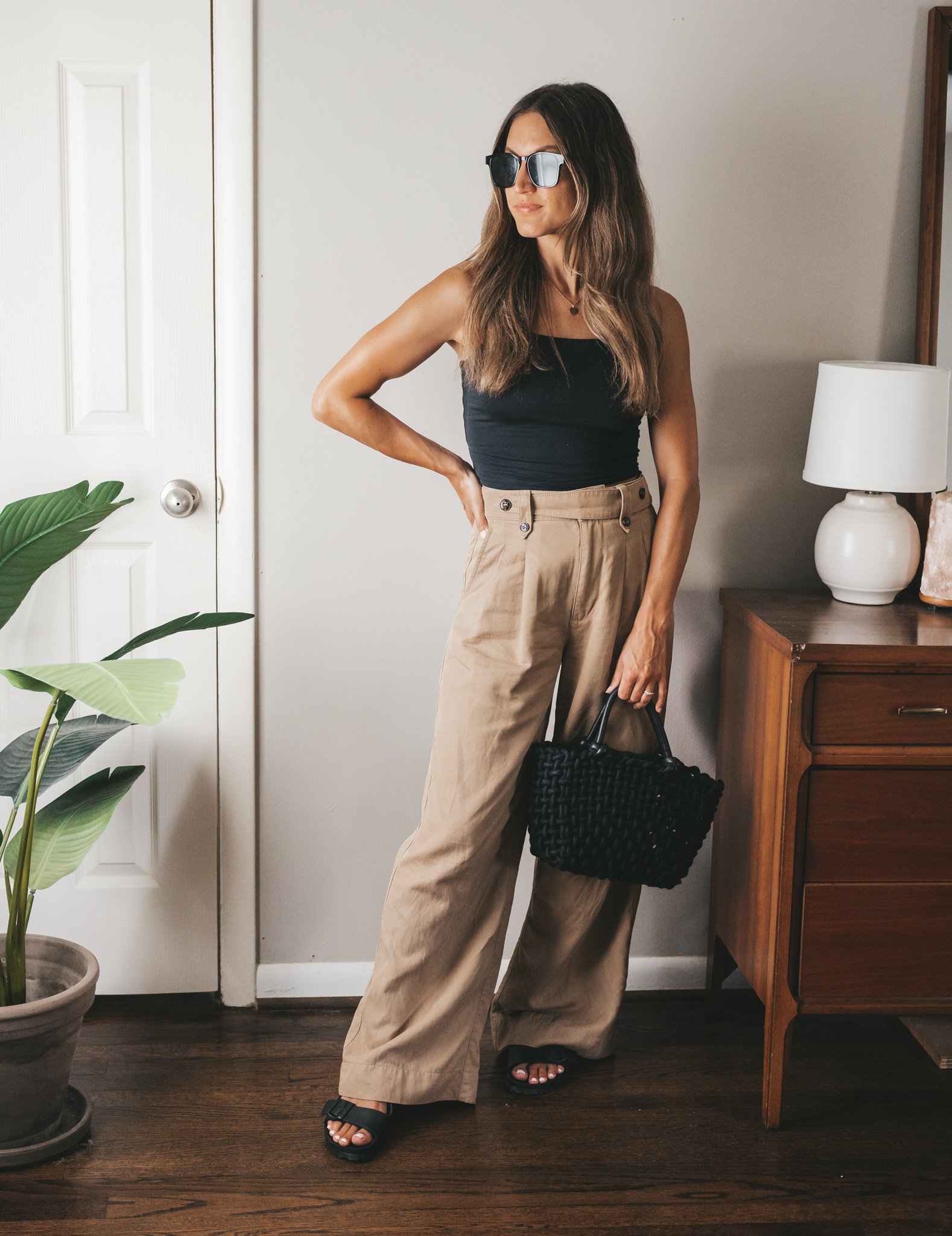 ashley wearing a black cropped spaghetti strap tank, wide leg tan trousers, black plastic birkenstocks, and a black woven bag