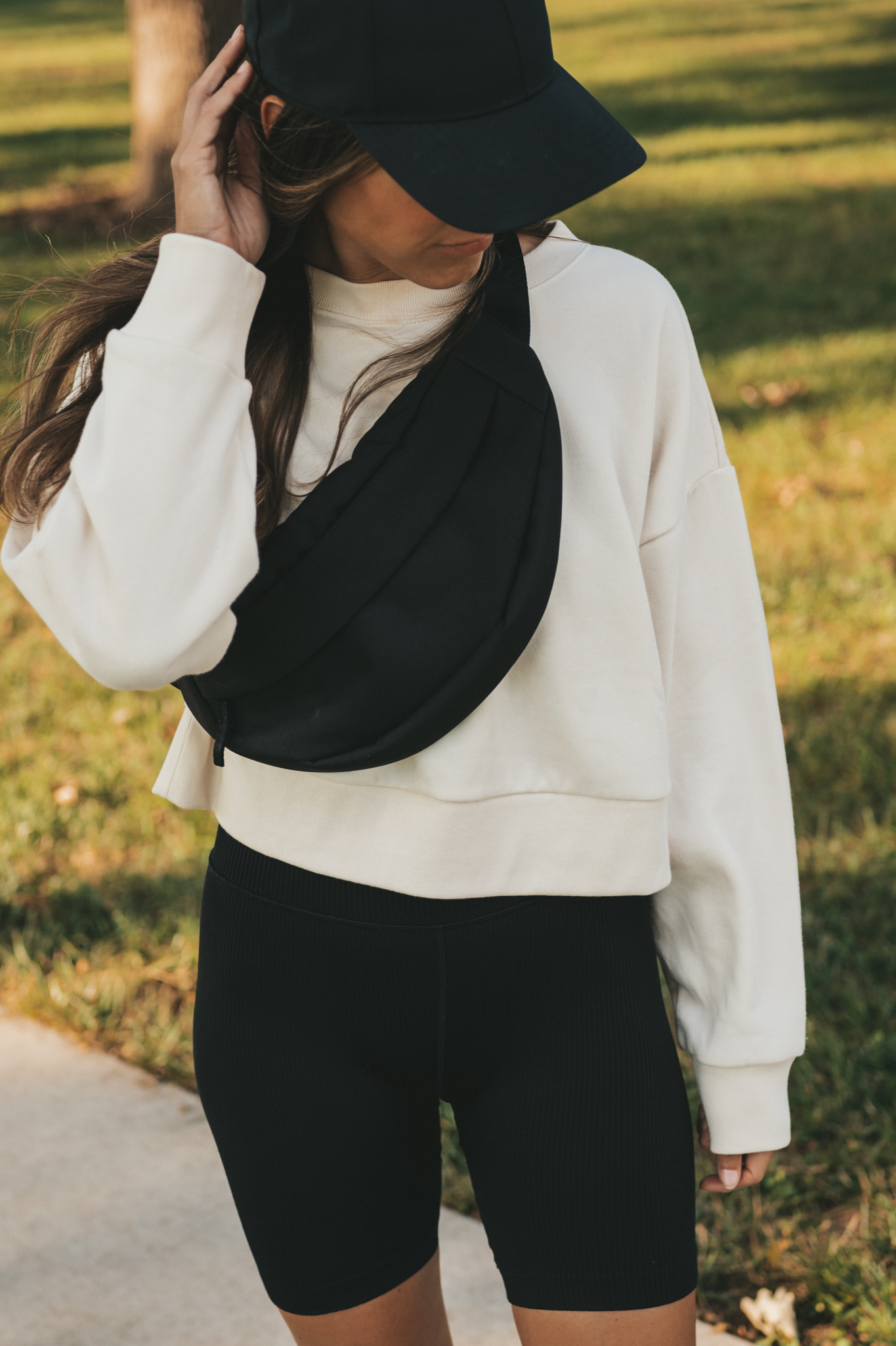 ashley wearing a cream colored cropped tank, black ribbed biker shorts, a black belt bag, and a black baseball cap