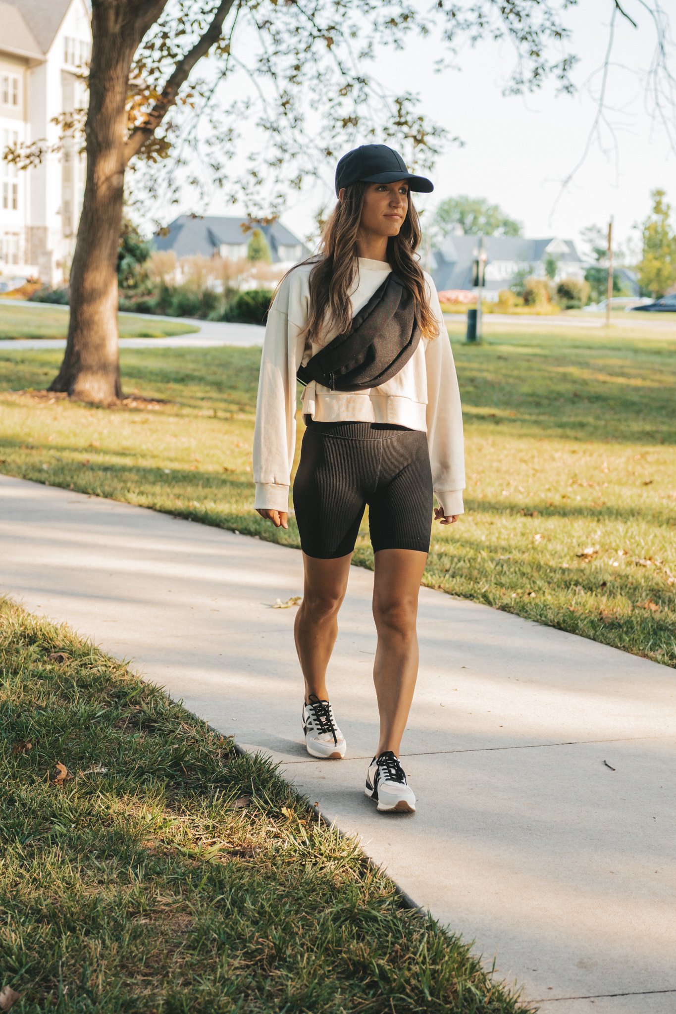 ashley wearing a cream colored cropped tank, black ribbed biker shorts, a black belt bag, and a black baseball cap