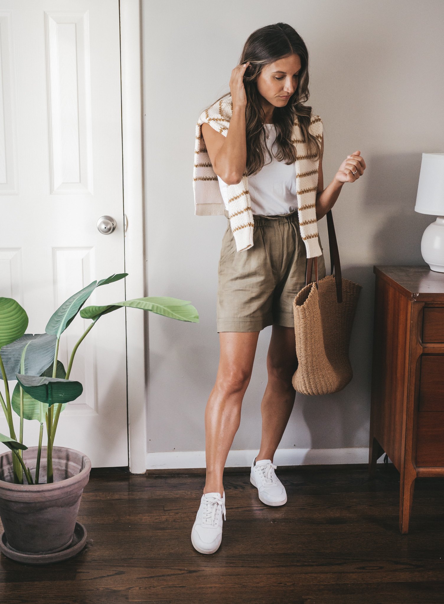 ashley wearing a white tank with a striped sweater worn over the shoulders, olive green paperbag shorts, white sneakers and a woven tote bag