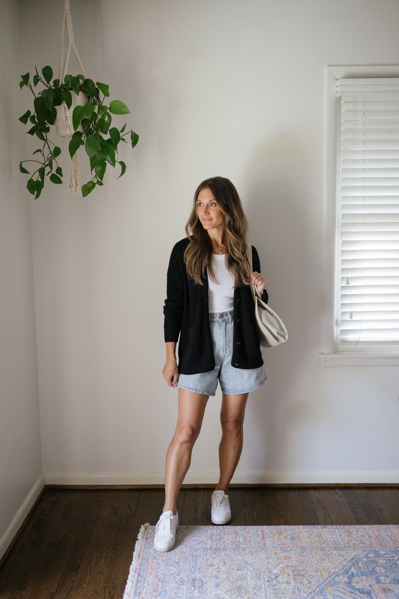 ashley wearing a white ribbed tank under a black cardigan with light wash denim shorts, white sneakers, and gray shoulder bag