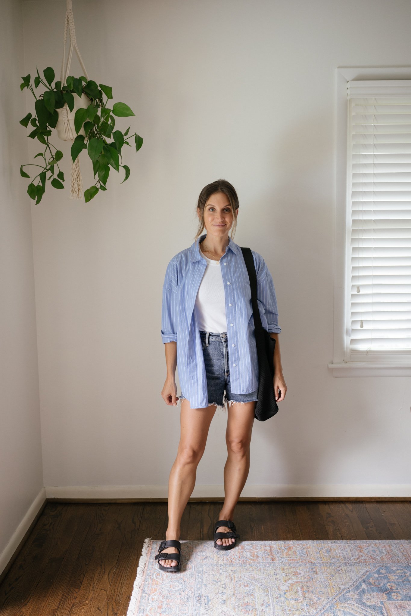 ashley wearing a white tank, oversized blue and white striped button down, cut-off denim shorts, and black Birkenstocks
