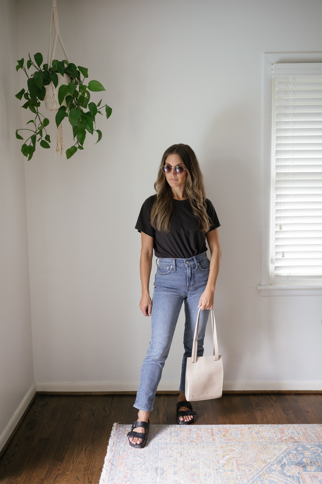 ashley wearing a black tee, mid-wash denim, black Birkenstocks, and a gray shoulder bag with round sunglasses