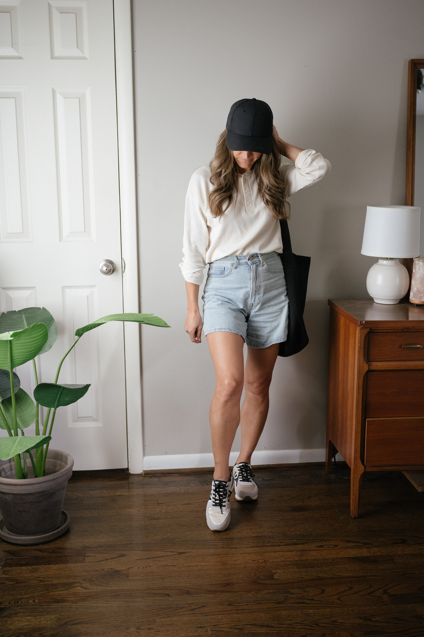 transitional outfit idea with a long sleeved cream colored waffle henley, denim shorts, casual sneakers, and a black baseball hat