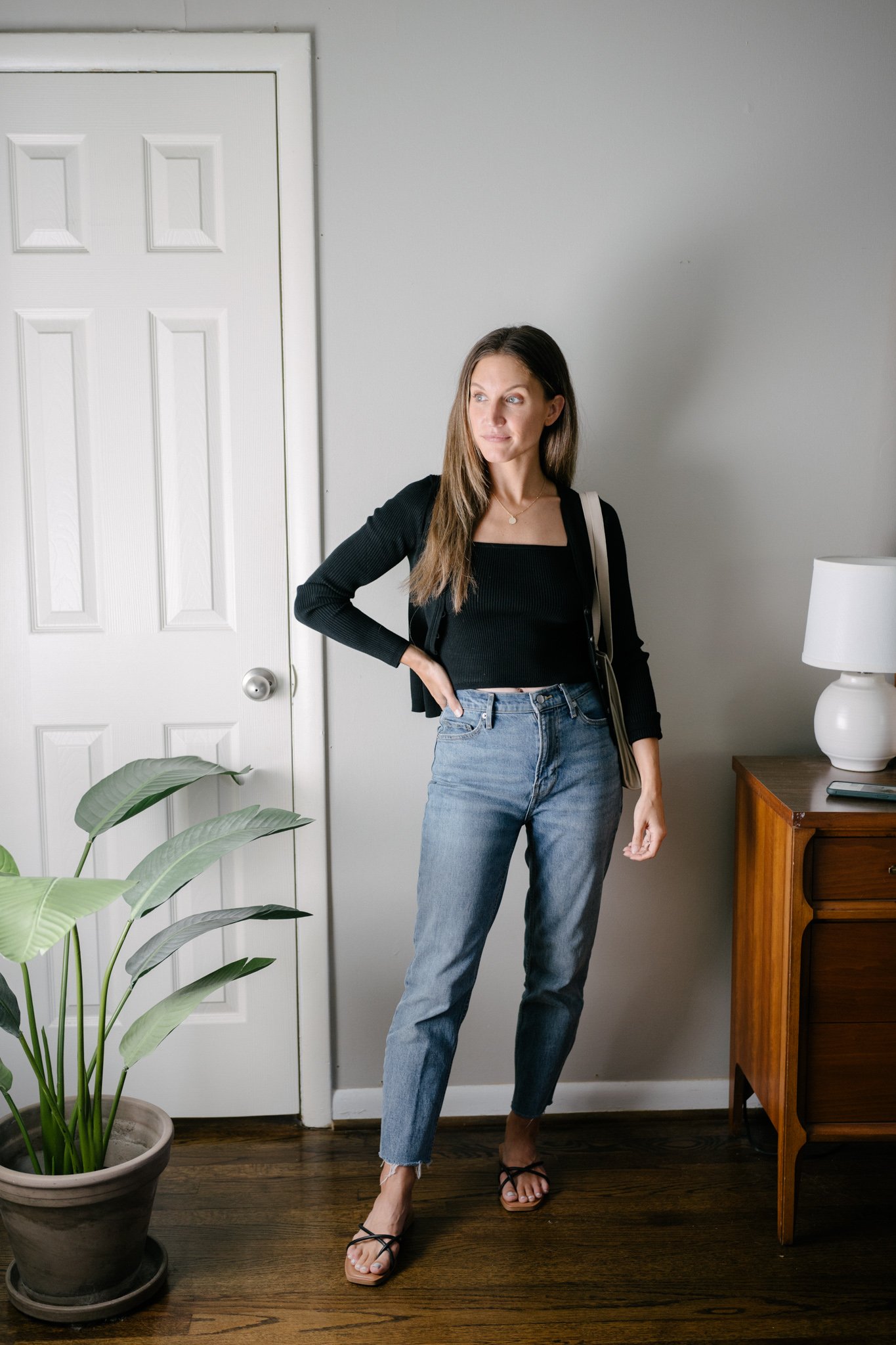 ashley wearing a black cropped cardigan over a black ribbed tank top, mid-wash mom jeans, and black strappy sandals with a gray shoulder bag