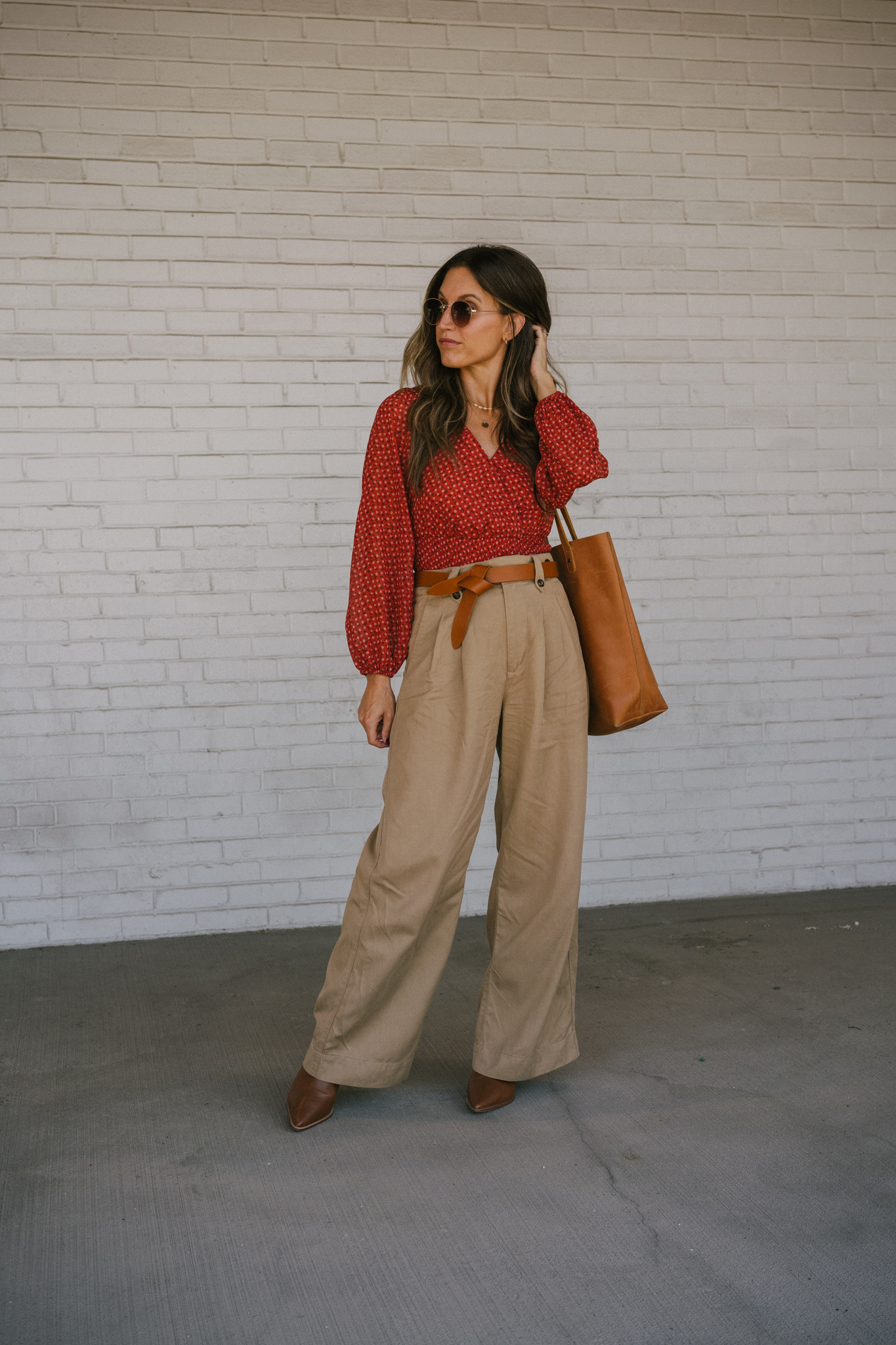 ashley wearing a fall floral blouse, wide leg trousers, a brown knotted belt, brown heeled ankle boots, and a brown leather tote bag with round gold frame sunglasses