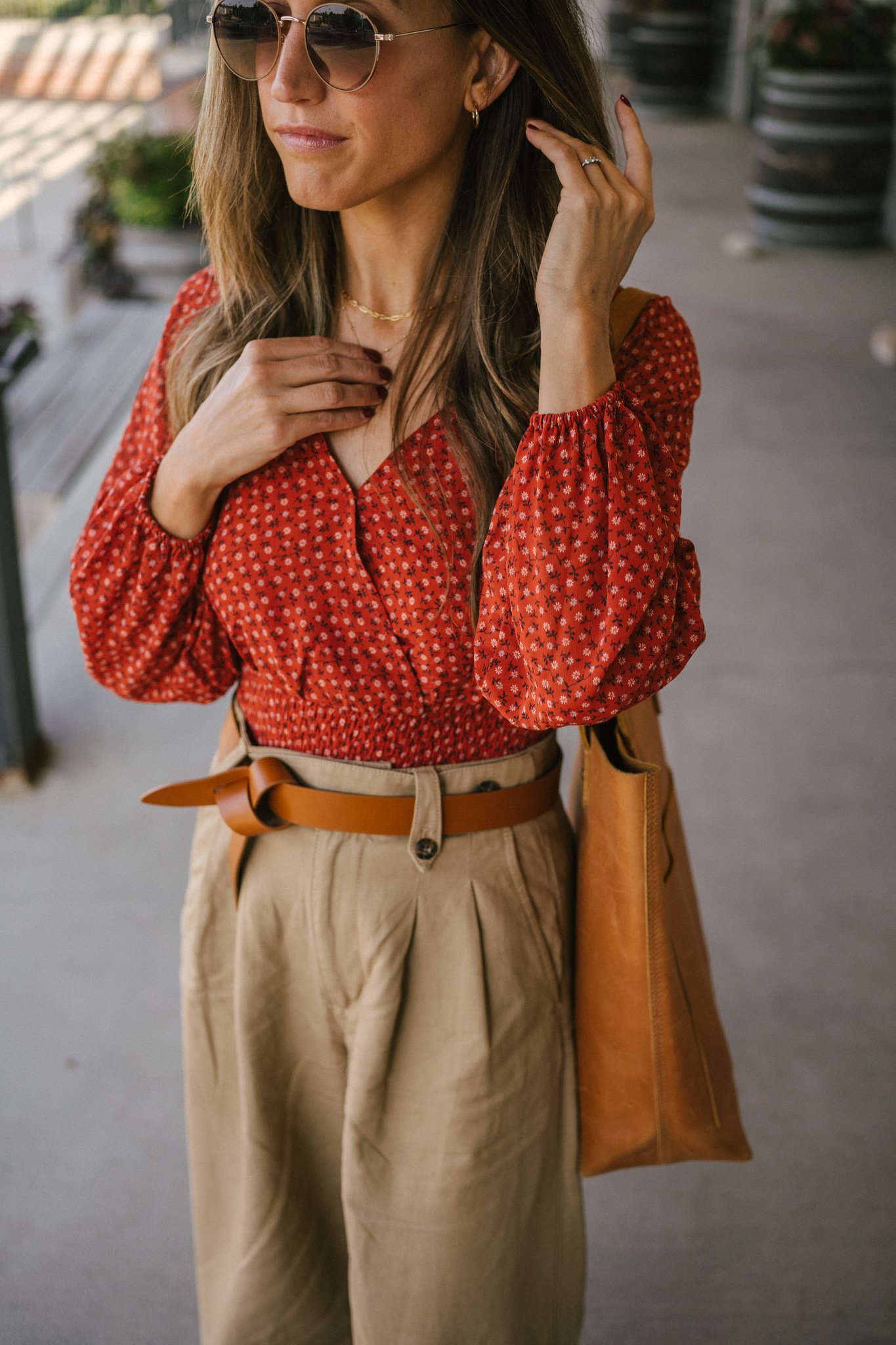 ashley wearing a fall floral blouse, wide leg trousers, a brown knotted belt, brown heeled ankle boots, and a brown leather tote bag with round gold frame sunglasses
