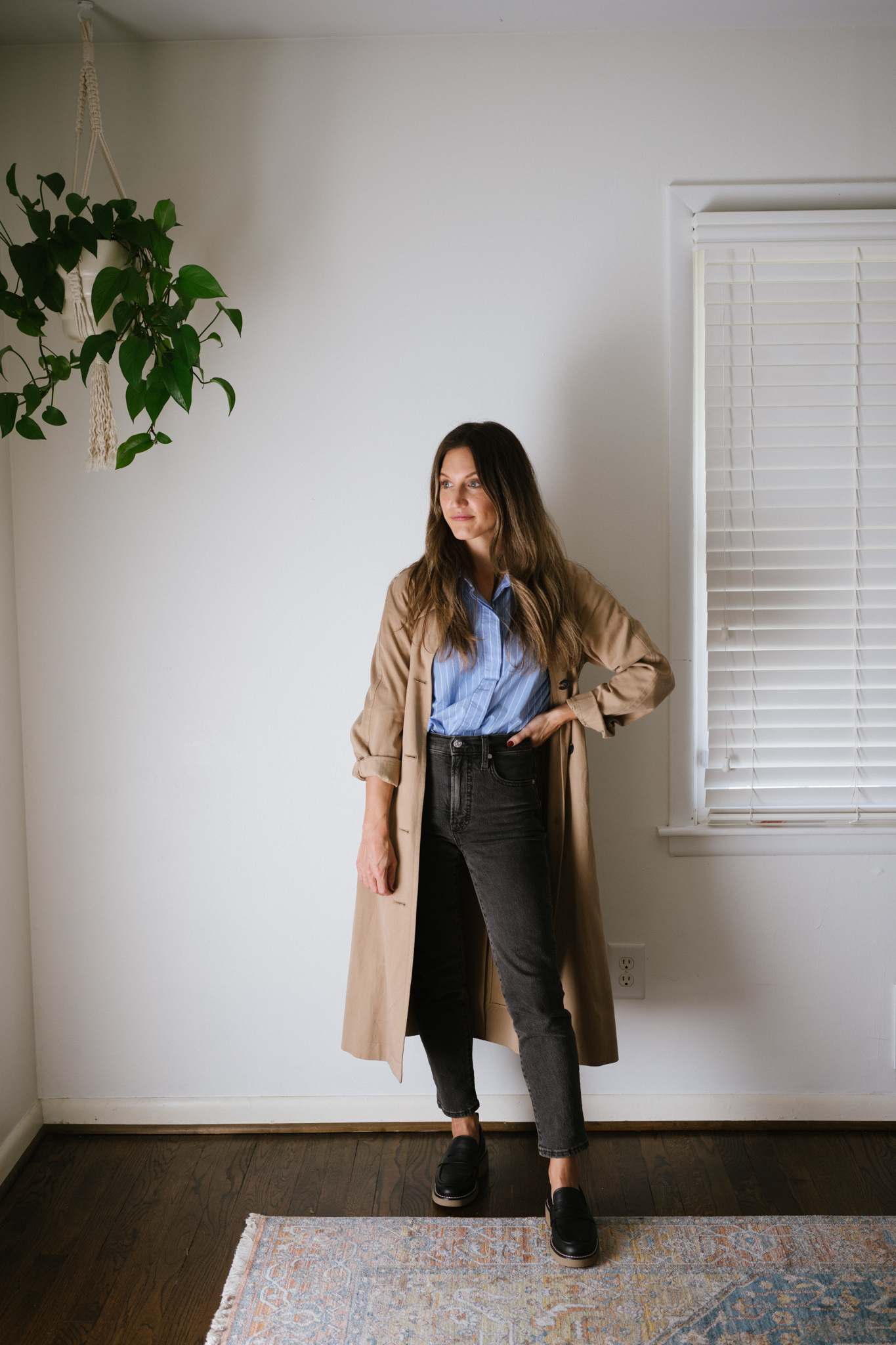 a minimalist fall outfit with a blue striped button down, black denim, tan trench coat, and black loafers