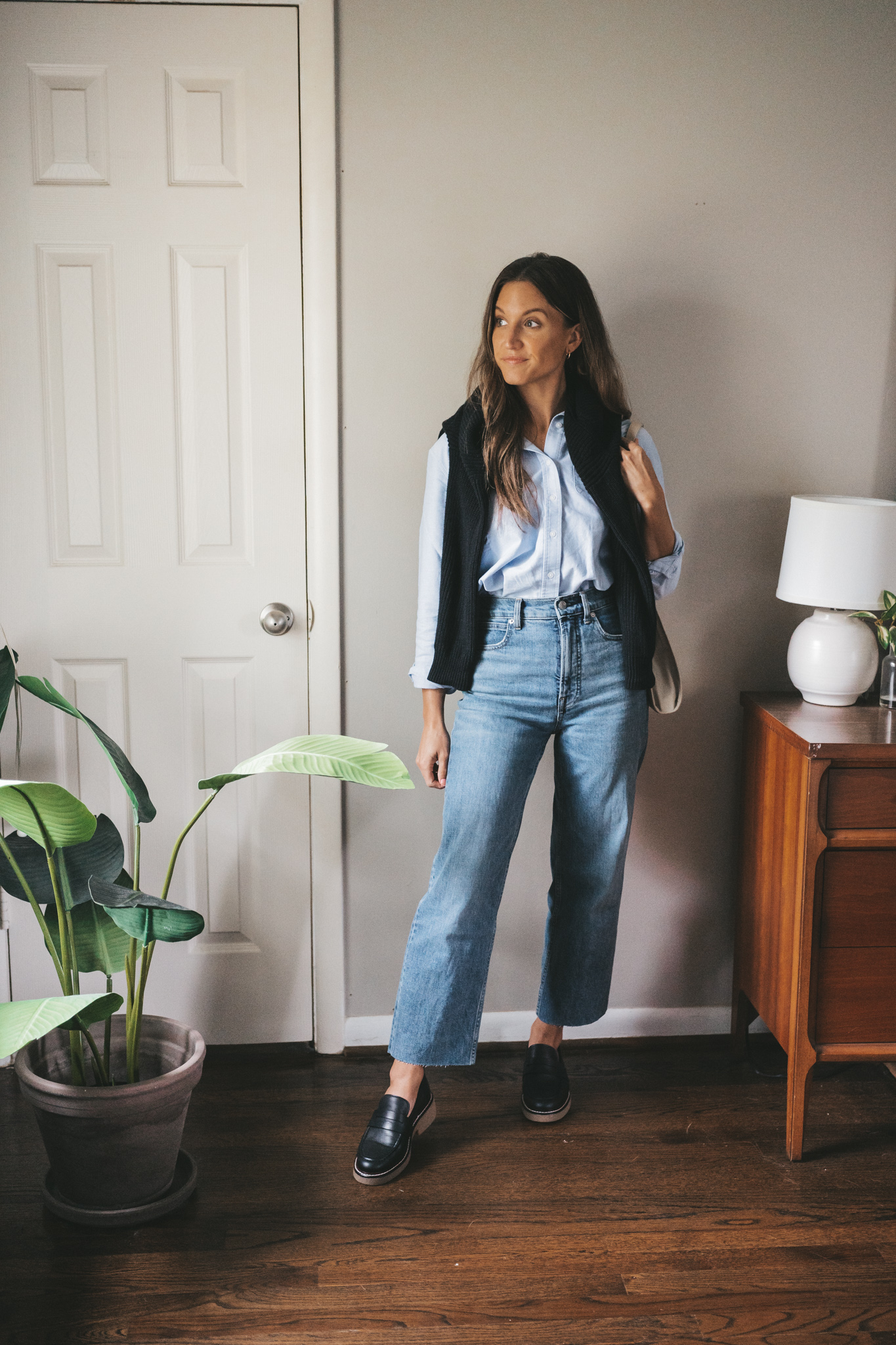 an early fall outfit with a light blue button down, black sweater worn over the shoulders, mid-wash straight denim, and black chunky oxfords