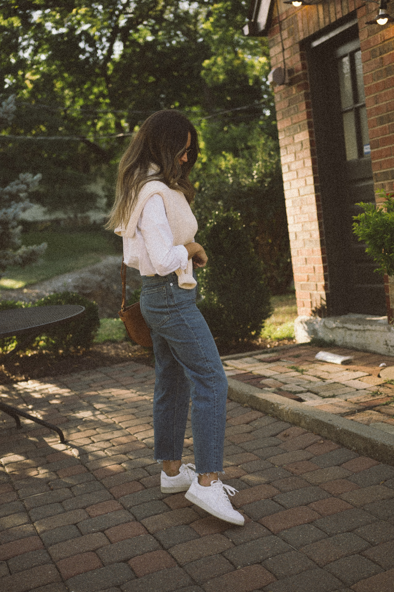 ashley wearing a white button down, cream sweater over the shoulders, and straight leg denim standing in front of a brick building with red pavers