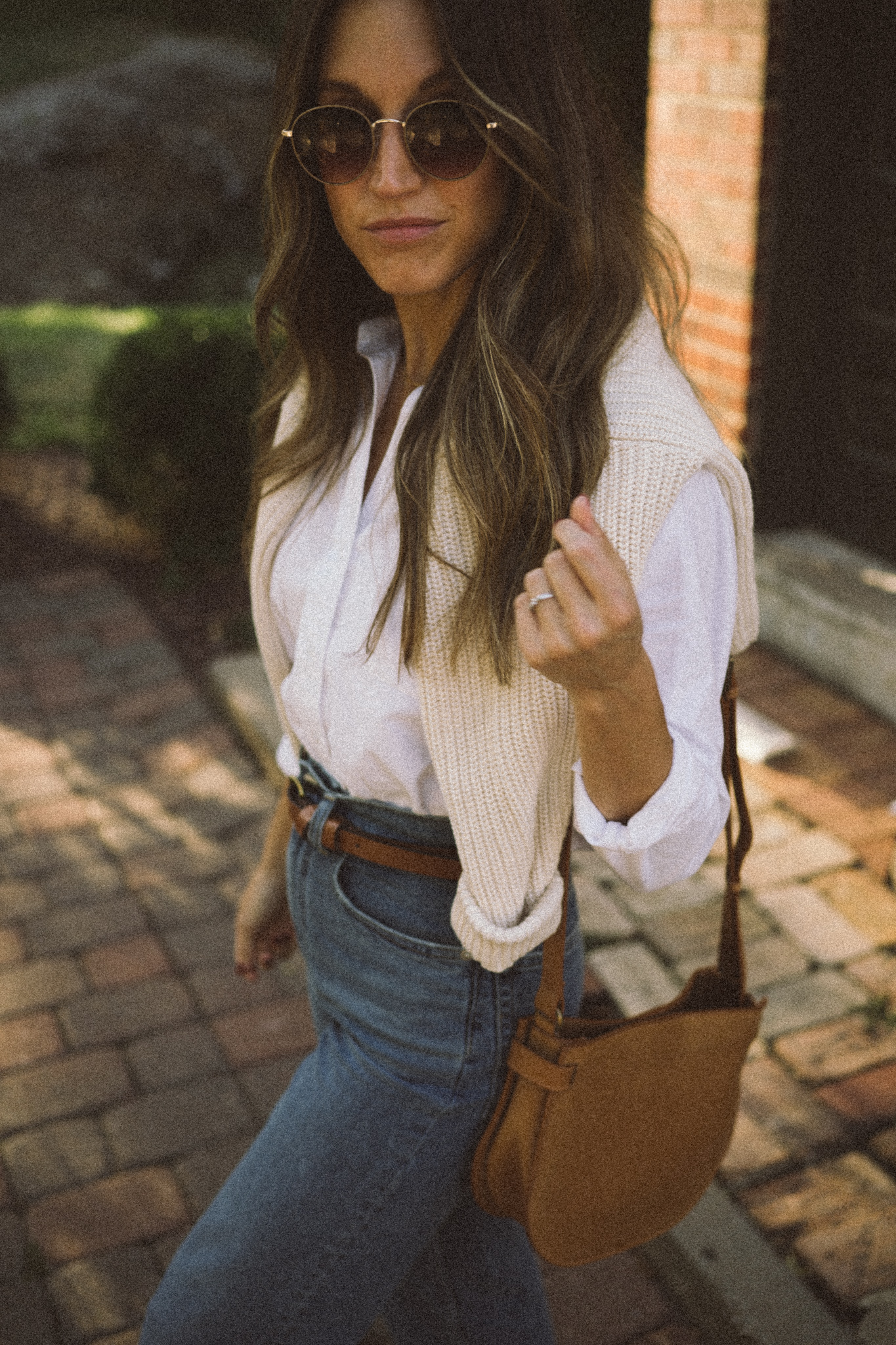 ashley wearing a white button down, cream sweater over the shoulders, and straight leg denim standing in front of a brick building with red pavers