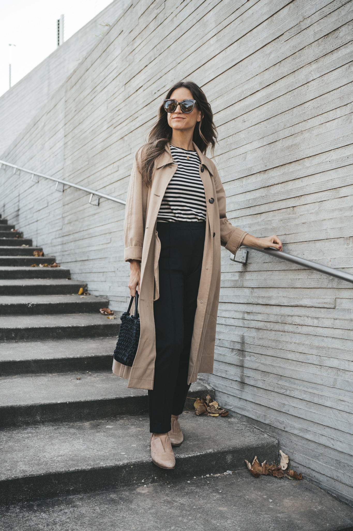ashley wearing the Everlane Dream pant paired with a striped tee, tan trench coat, nude Oxfords, and a black woven bag