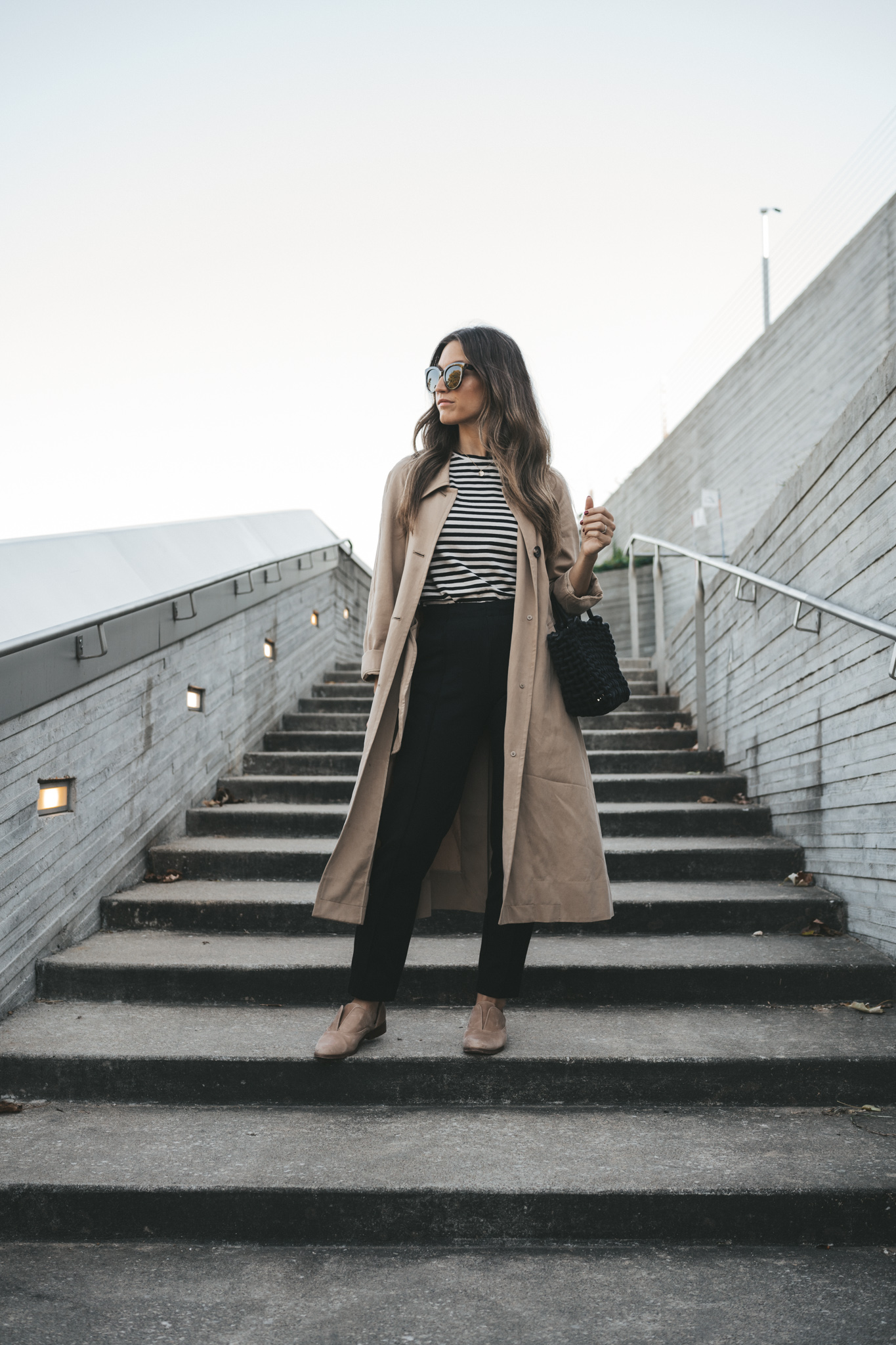 ashley wearing the Everlane Dream pant paired with a striped tee, tan trench coat, nude Oxfords, and a black woven bag