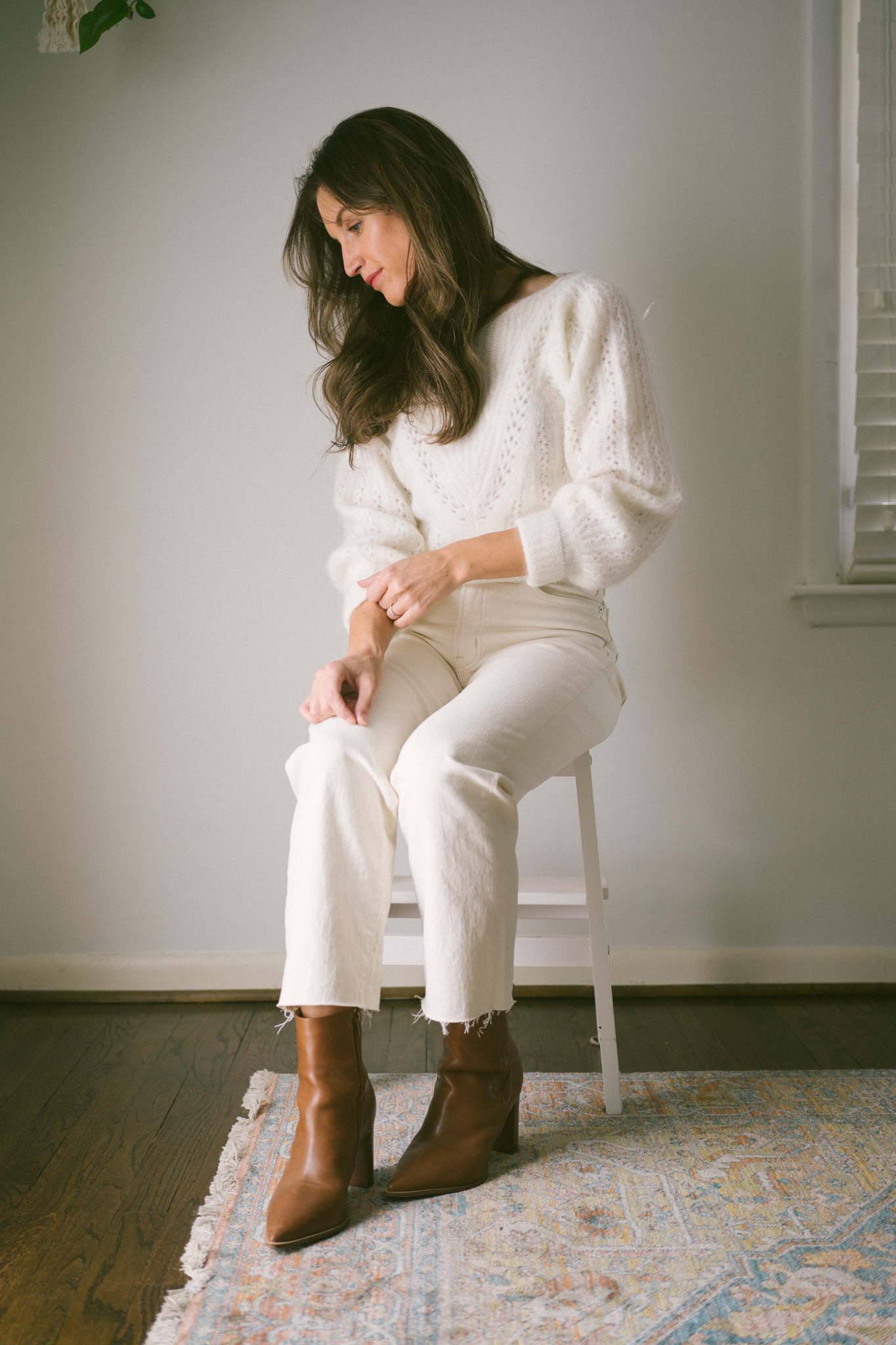 ashley wearing a white sweater, white denim, and brown boots sitting on a stool