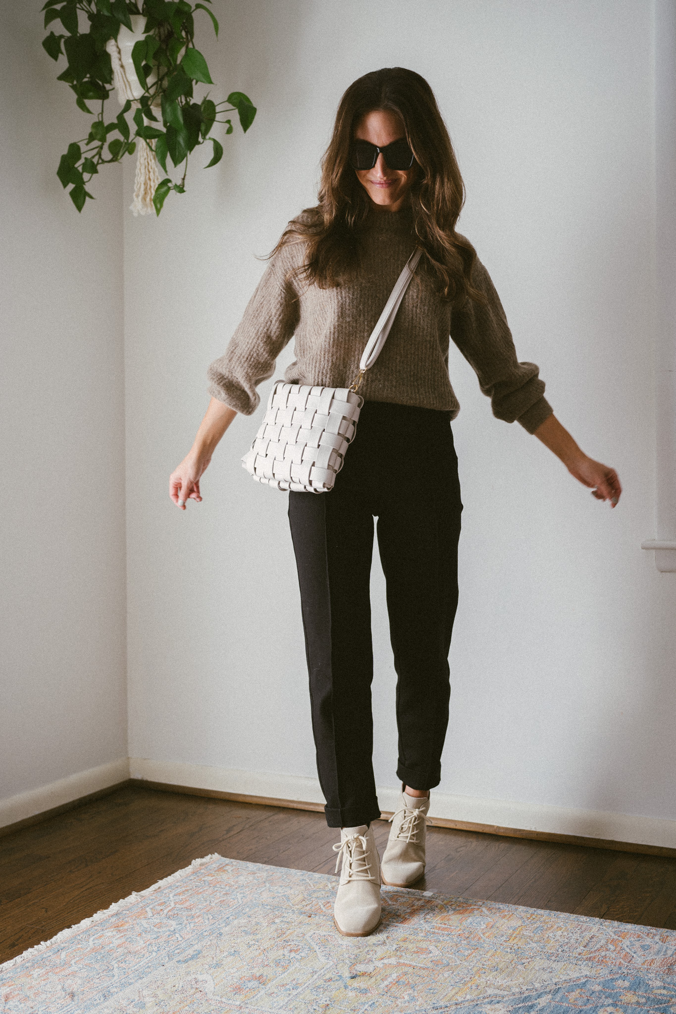 ashley wearing a taupe oversized sweater, black pants, and bone colored boots with laces with a woven bone colored bag and black sunglasses