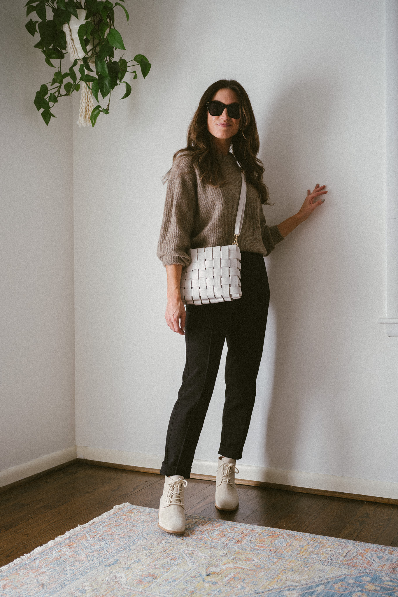 ashley wearing a taupe oversized sweater, black pants, and bone colored boots with laces with a woven bone colored bag and black sunglasses