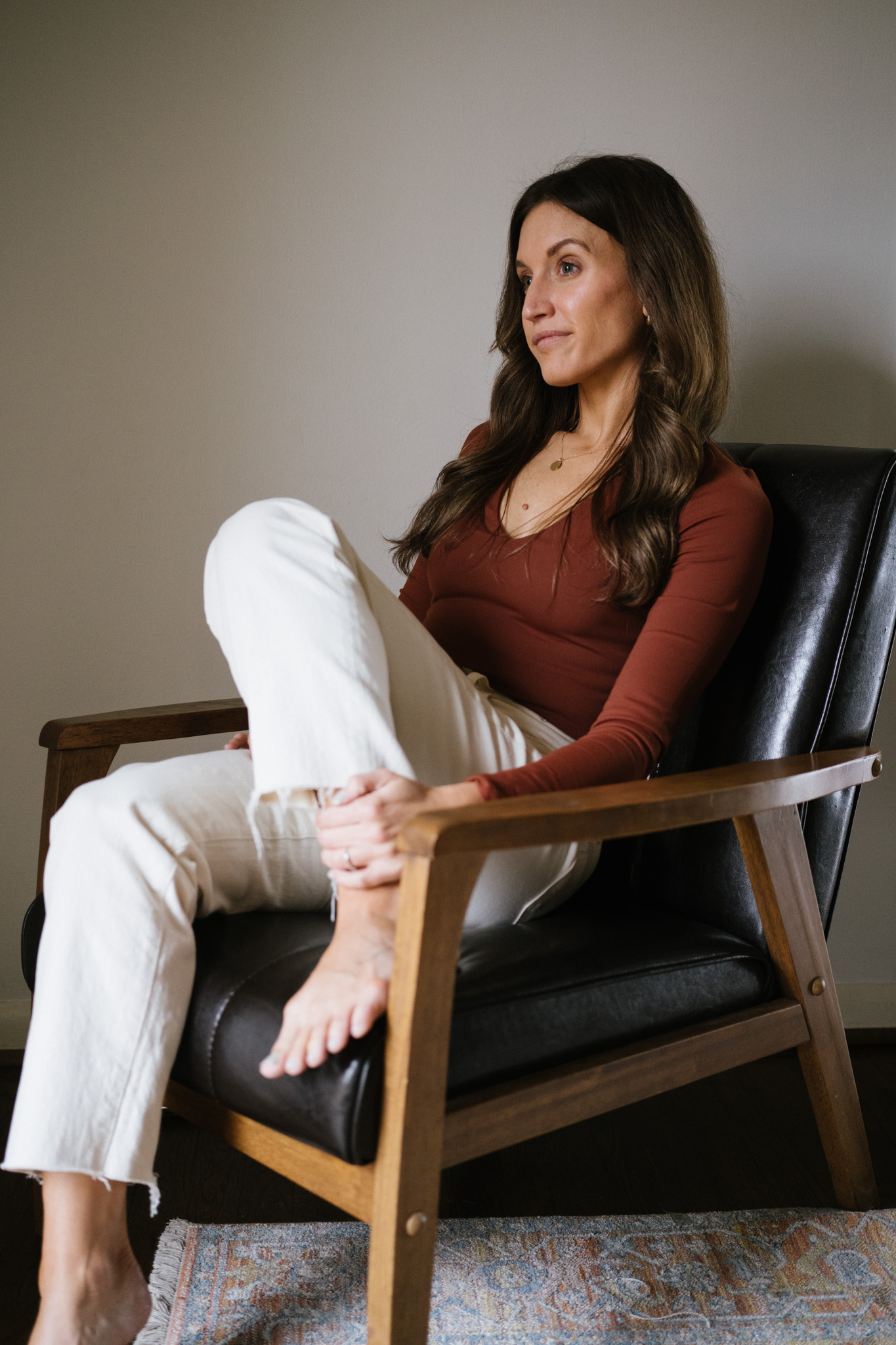 ashley wearing a reddish brown bodysuit and white jeans sitting in a black leather chair