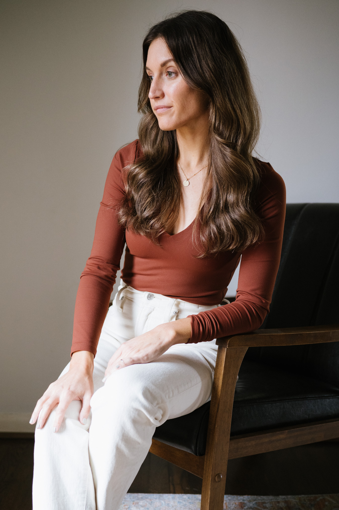 ashley wearing a reddish brown bodysuit and white jeans sitting in a black leather chair