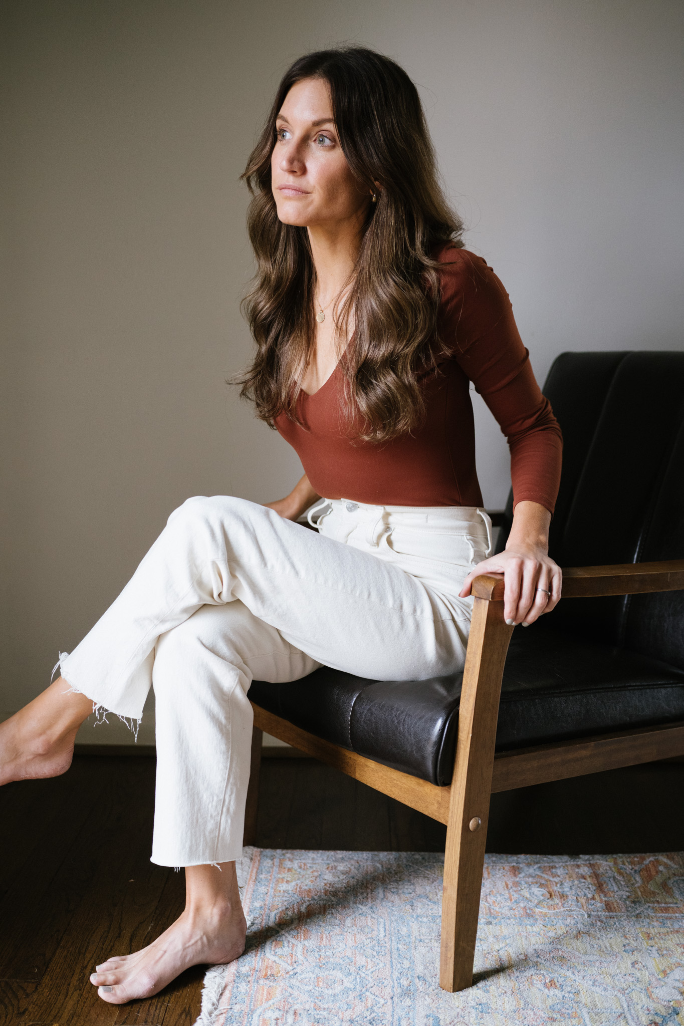 ashley wearing a reddish brown bodysuit and white jeans sitting in a black leather chair