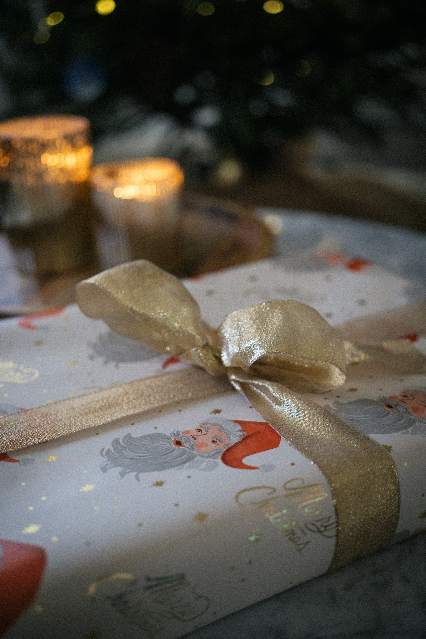 a wrapped christmas present with gold ribbon and lit candles and a christmas tree in the background