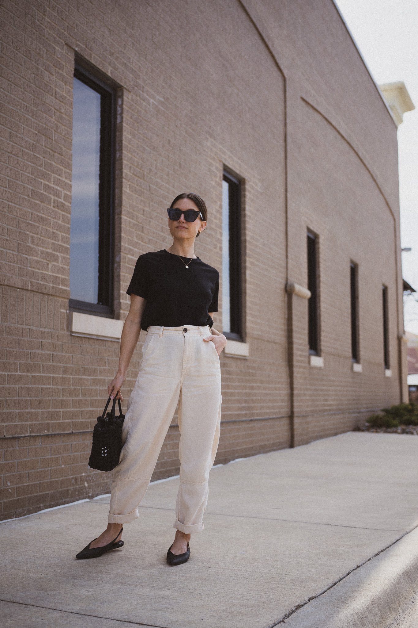 ashley wearing  a black tee, beige chino pants, and black slingback flats standing next to a gray building