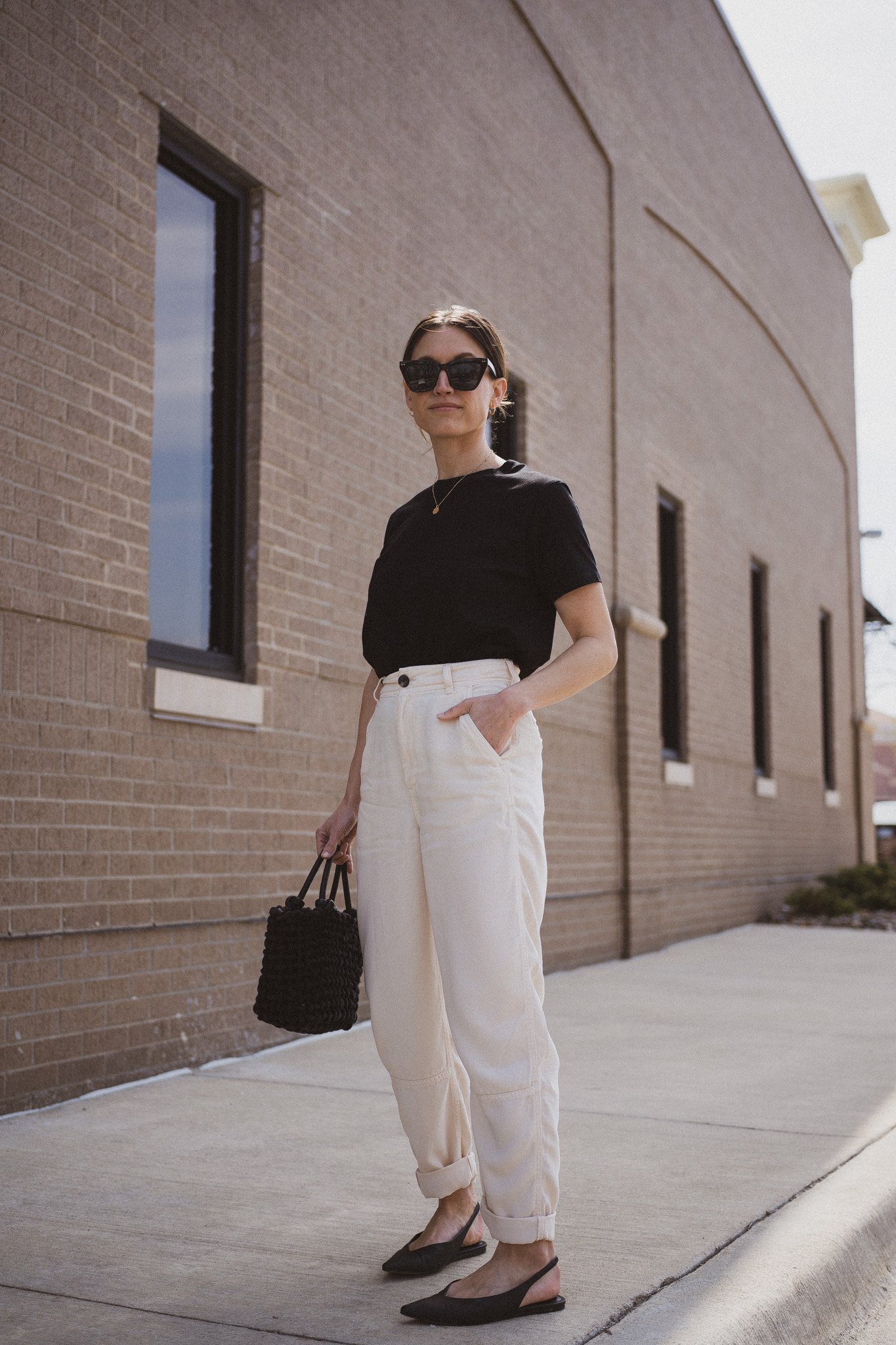 ashley wearing  a black tee, beige chino pants, and black slingback flats standing next to a gray building