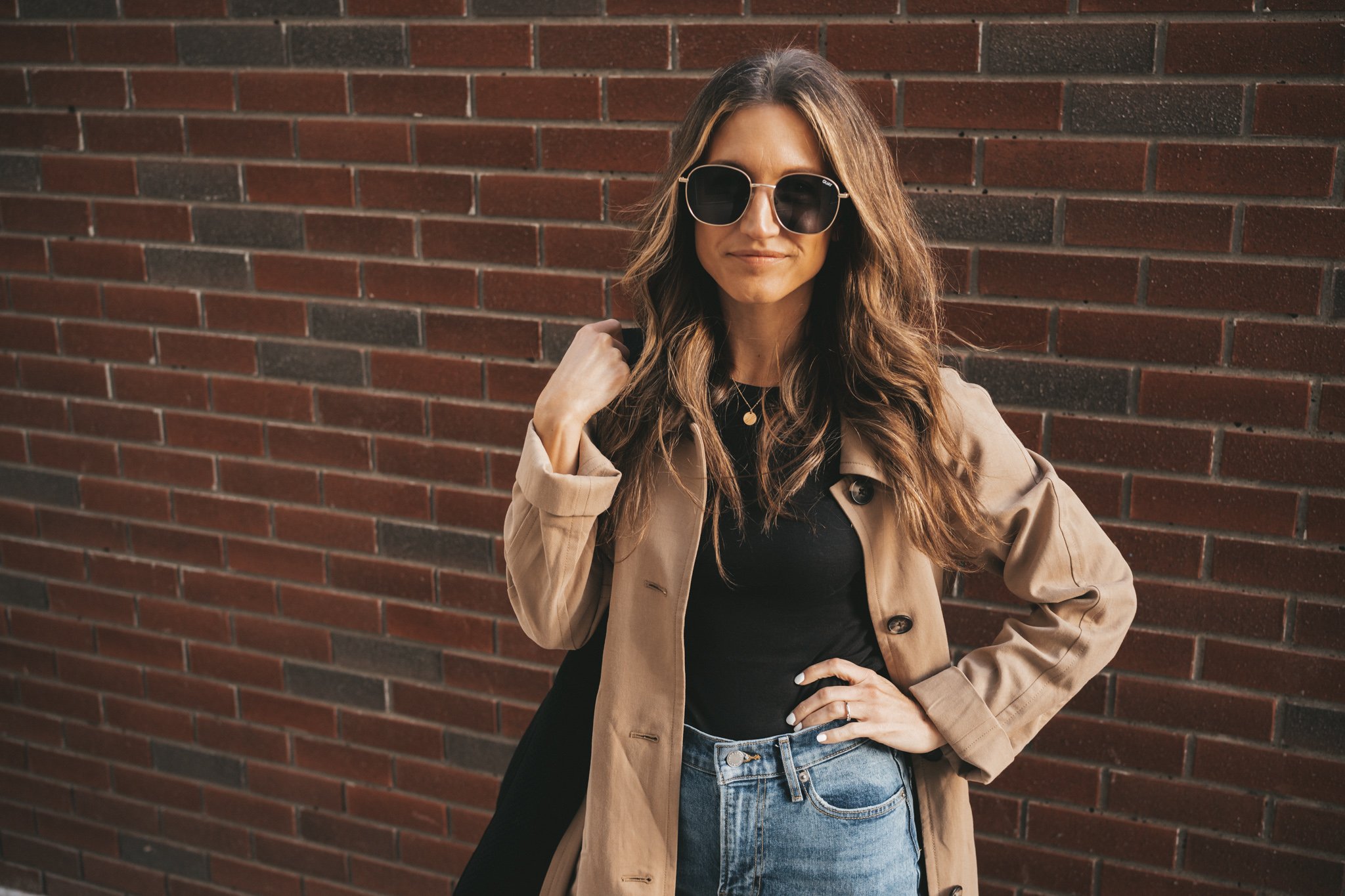 ashley wearing a tan trench coat, denim, and white sneakers standing in front of a brick wall. 