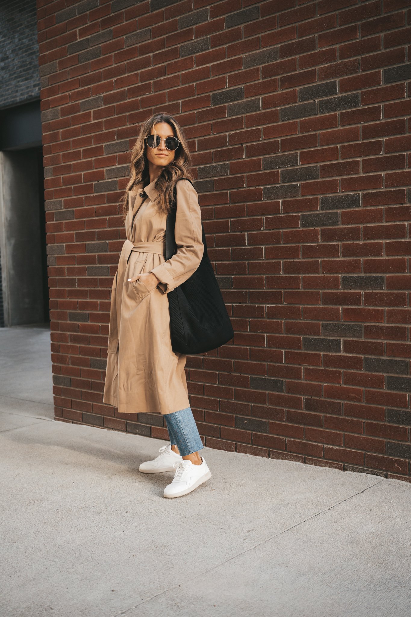 ashley wearing a tan trench coat, denim, and white sneakers standing in front of a brick wall. 