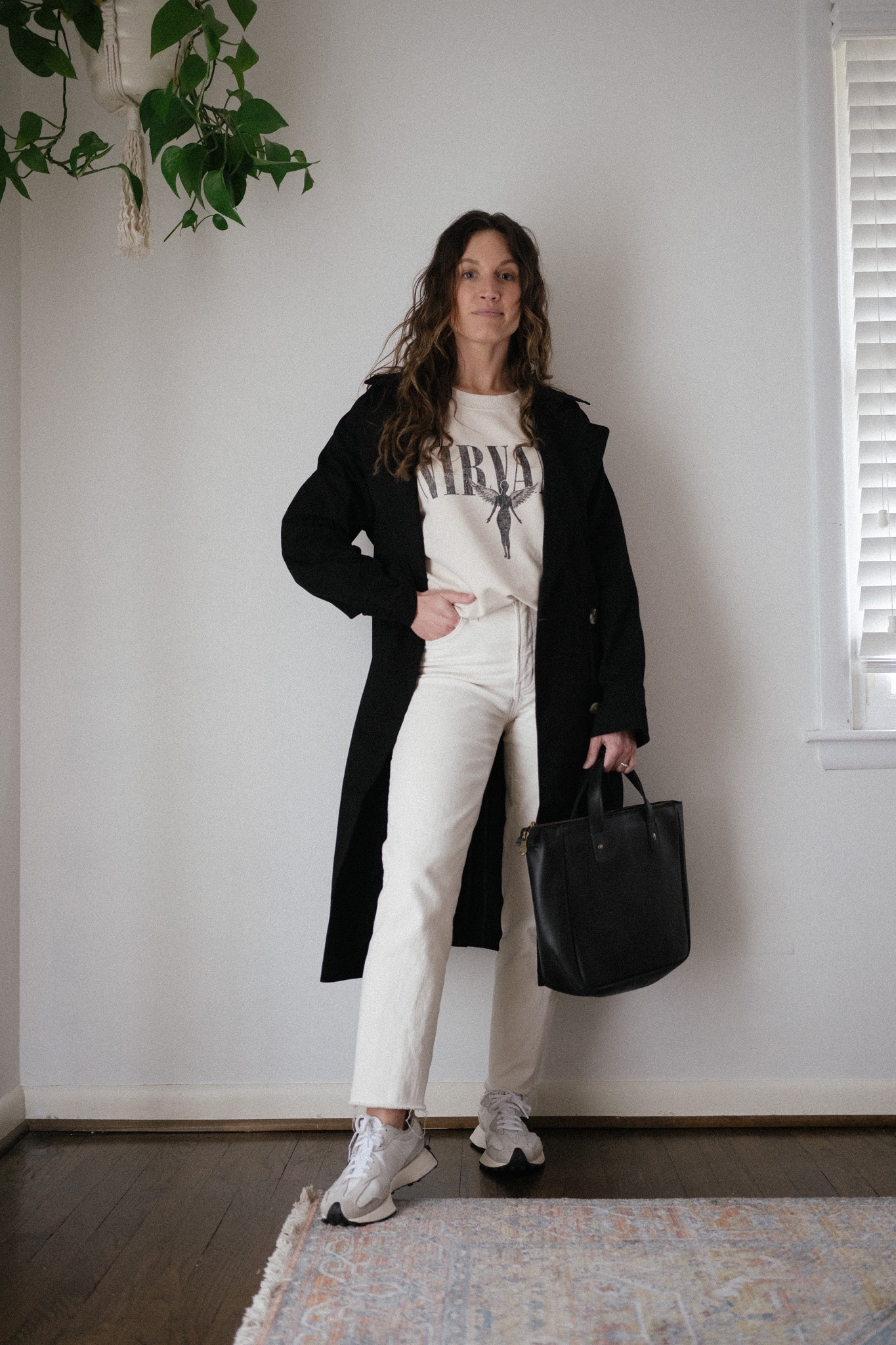 casual outfit idea with a beige graphic sweatshirt, black trench coat, white denim, and new balance sneakers