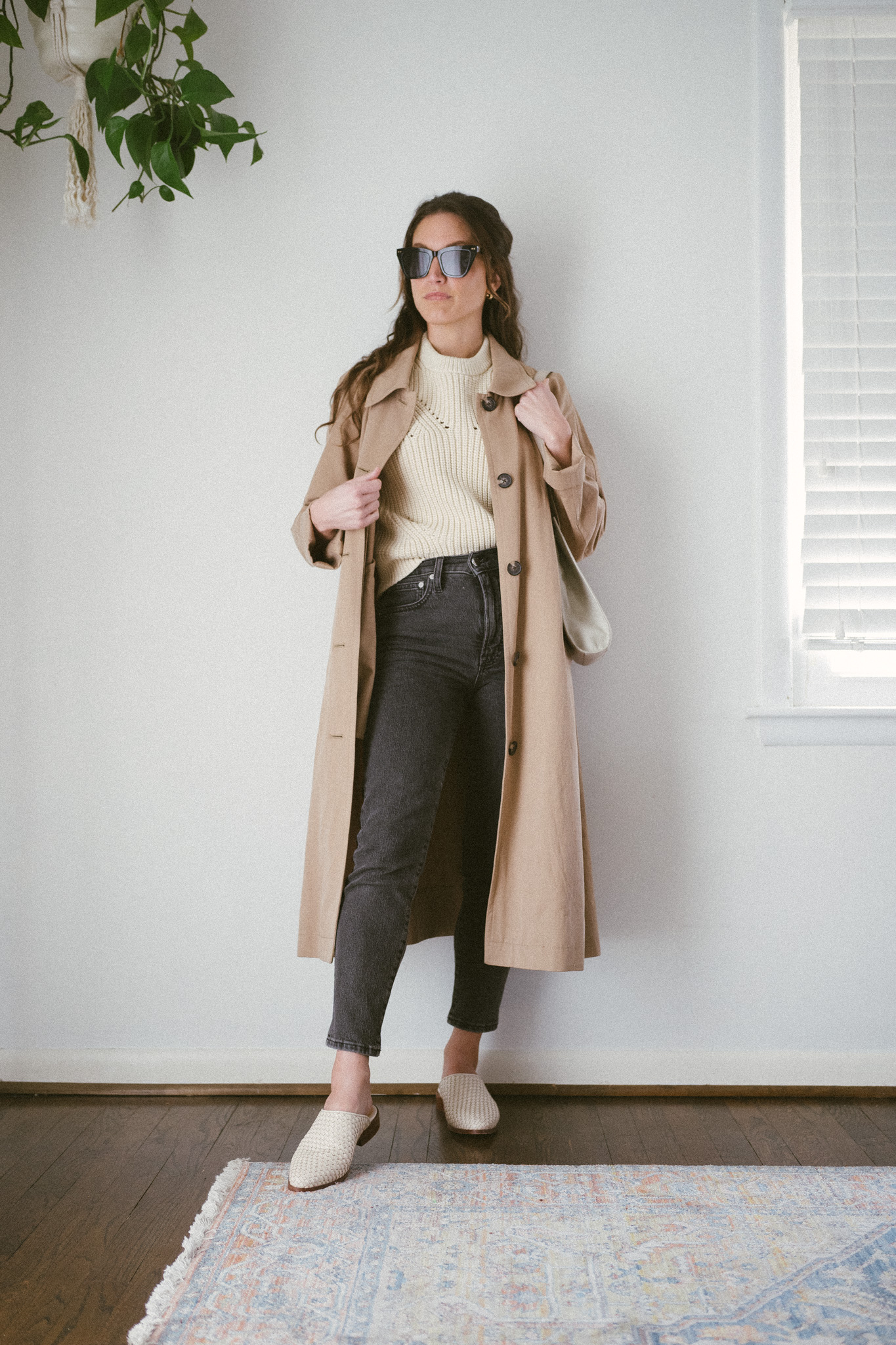 spring outfit idea with a cream colored sweater tee, tan trench coat, washed black denim, and white woven mules