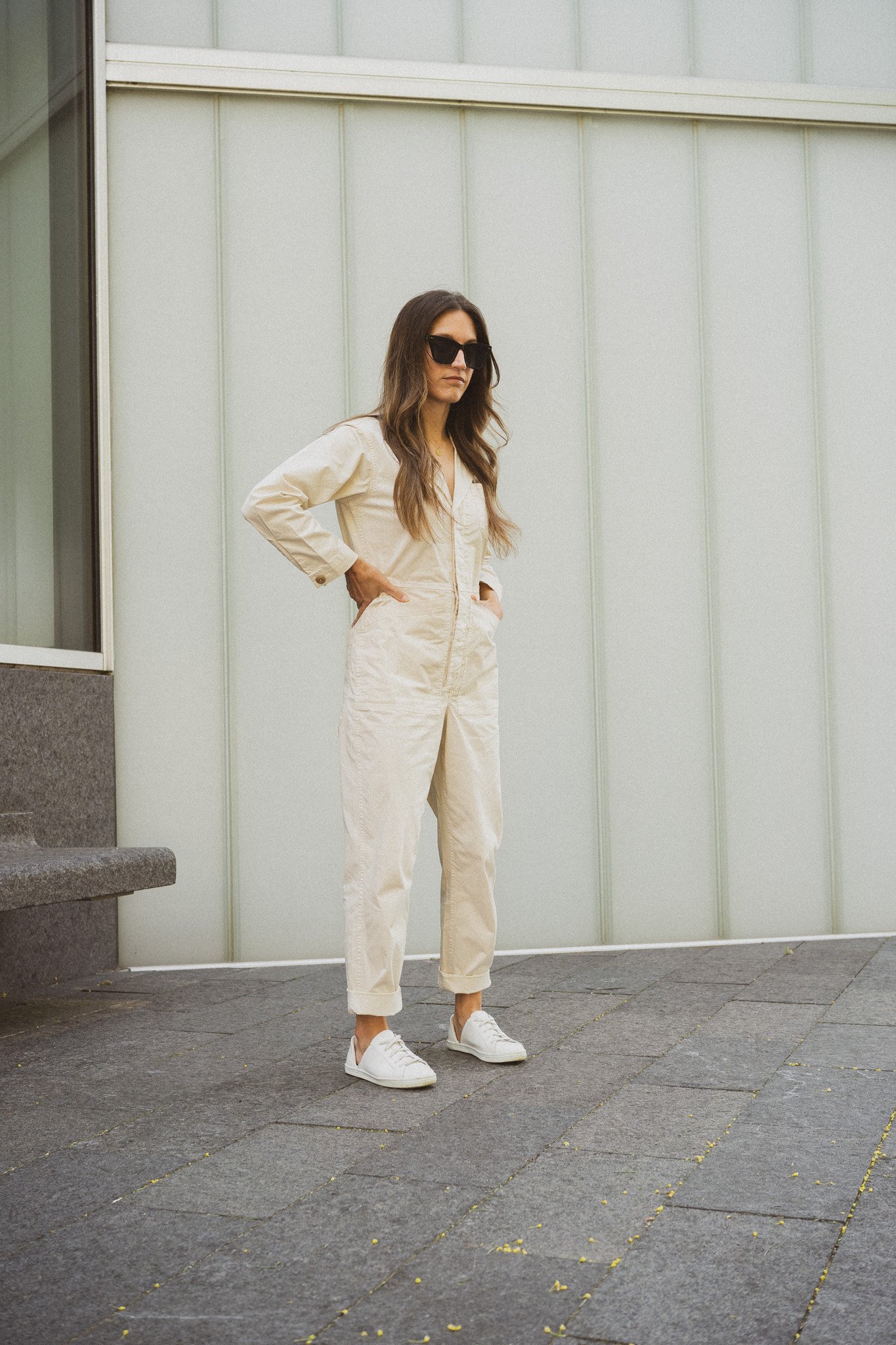 ashley wearing the alex mill standard jumpsuit in the oat milk color, white sneakers, and a green handbag