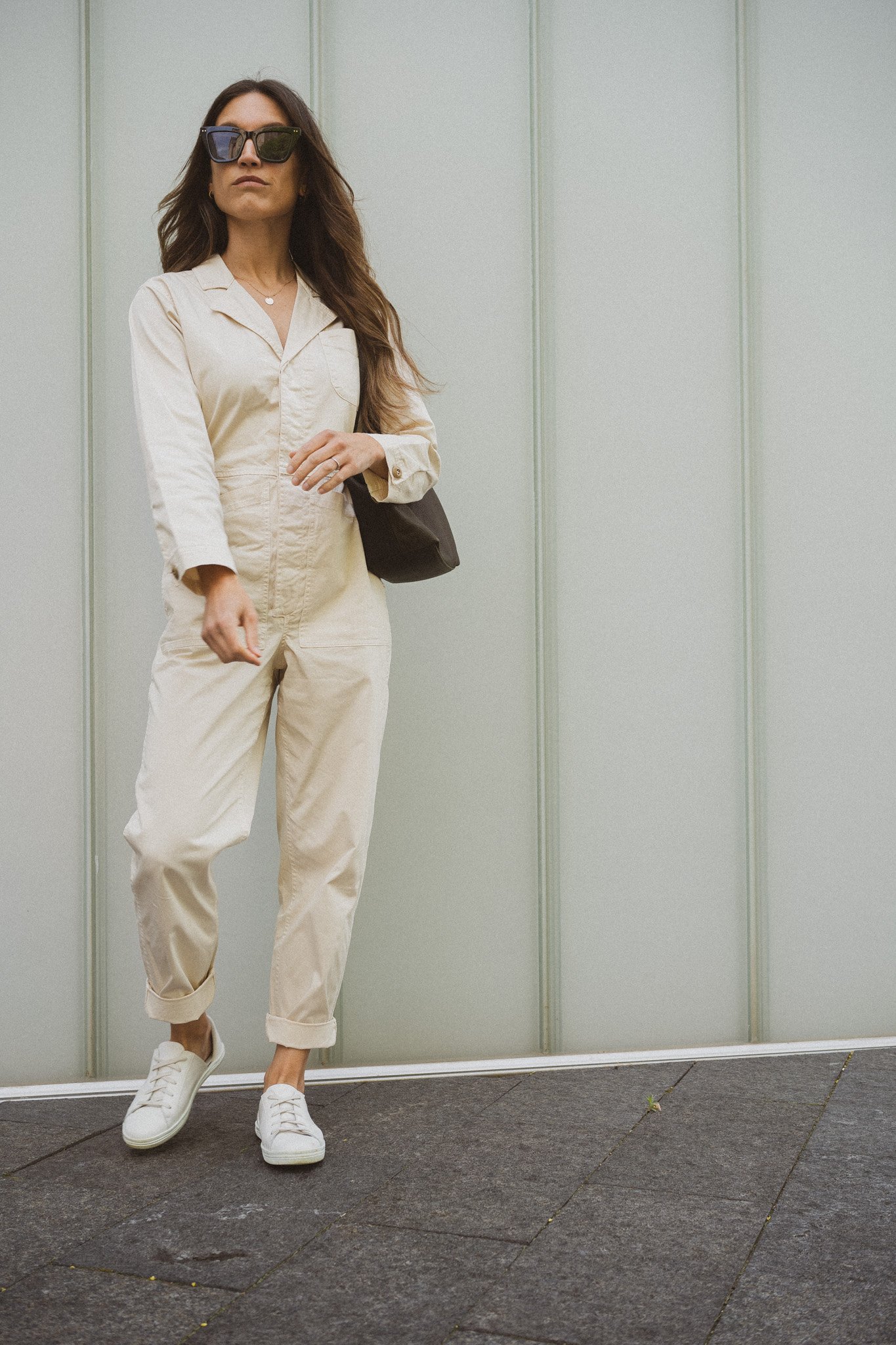 ashley wearing the alex mill standard jumpsuit in the oat milk color, white sneakers, and a green handbag