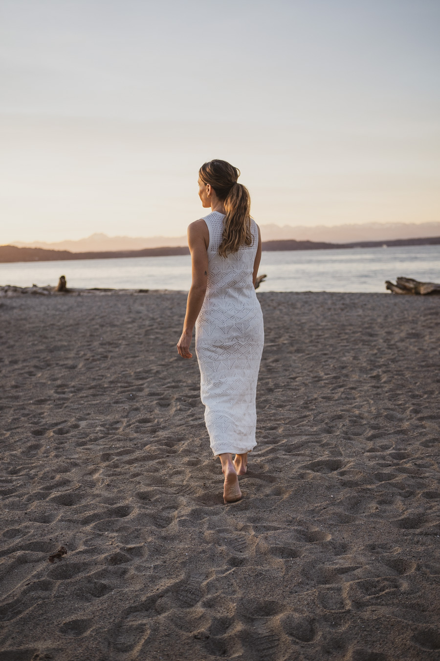 ashley on a beach wearing an embroidered sleeveless white dress and carrying sandals