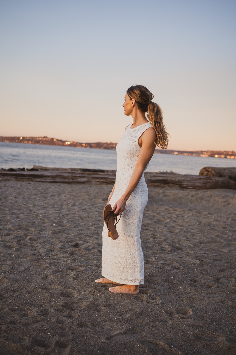 ashley on a beach wearing an embroidered sleeveless white dress and carrying sandals