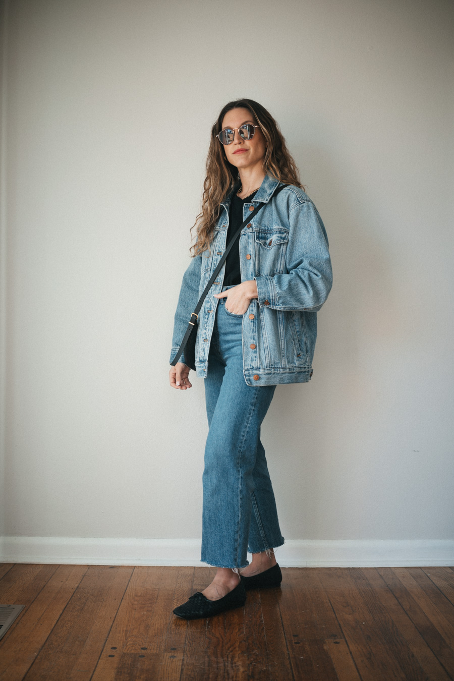 ashley wearing a black tee, oversized denim jacket, medium wash straight cropped jeans, and black woven ballet flats