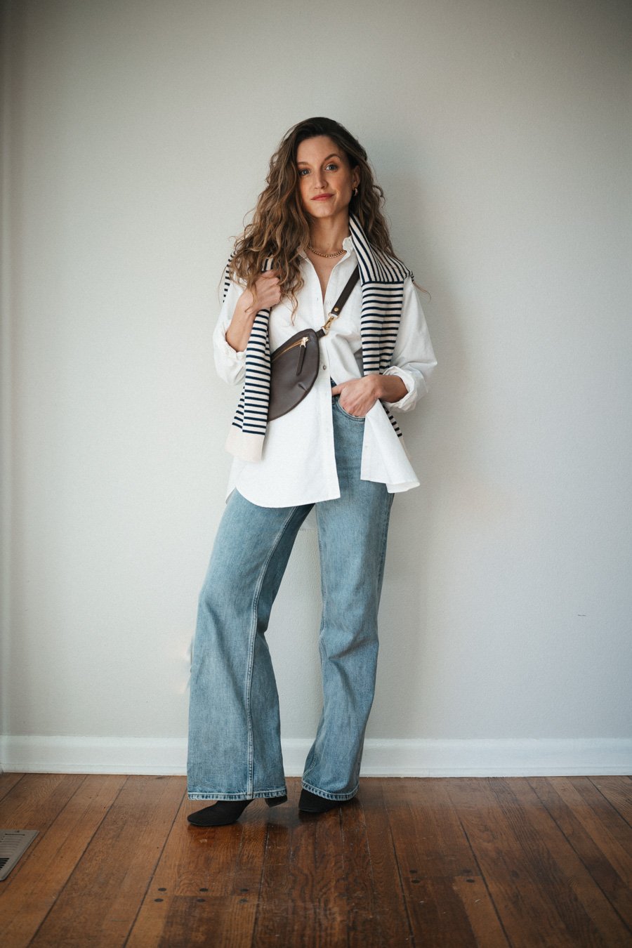 spring transitional outfit with an oversized white button down. light wash wide leg jeans, brown boots, brown sling bag, and a navy and ecru striped sweater worn over the shoulders