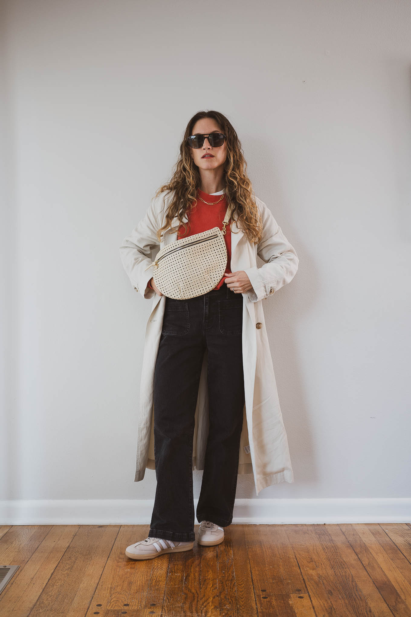 fall capsule outfit with a red sweatshirt, beige trench coat, black jeans, and tan Adidas Sambas with a cream sling bag