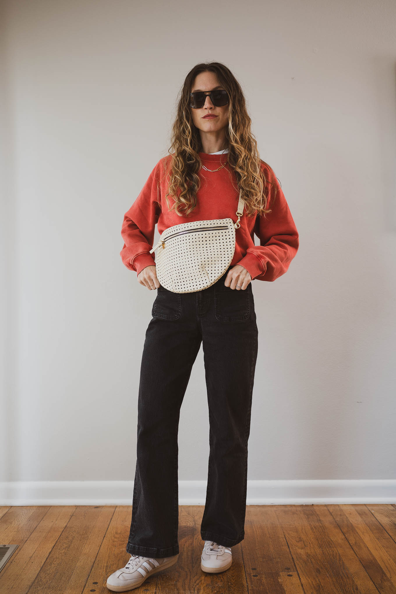 fall capsule outfit with a red sweatshirt, beige trench coat, black jeans, and tan Adidas Sambas with a cream sling bag