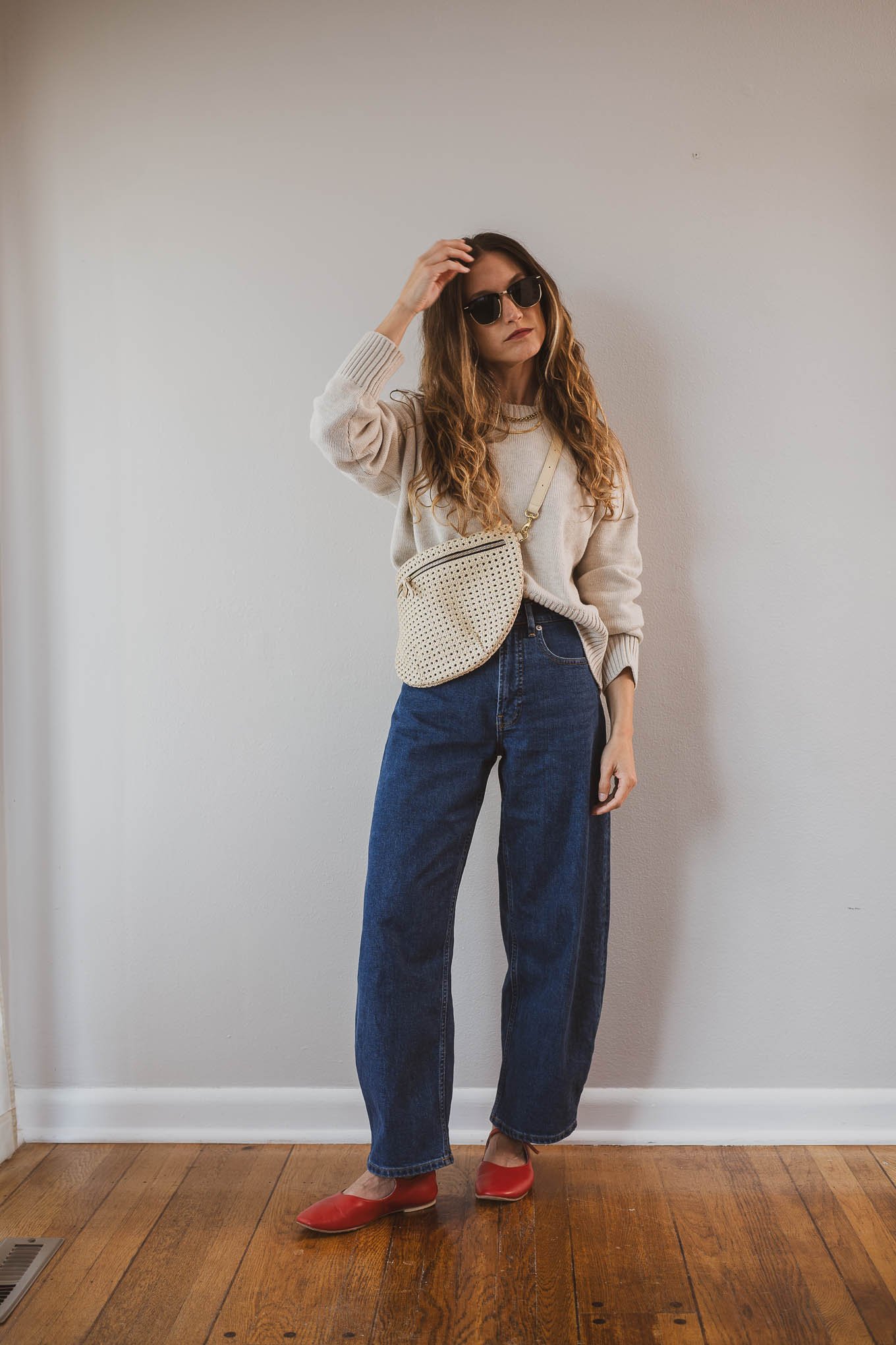 fall capsule wardrobe outfit with a beige cotton sweater, barrel jeans, red mary jane ballet flats, and a cream sling bag