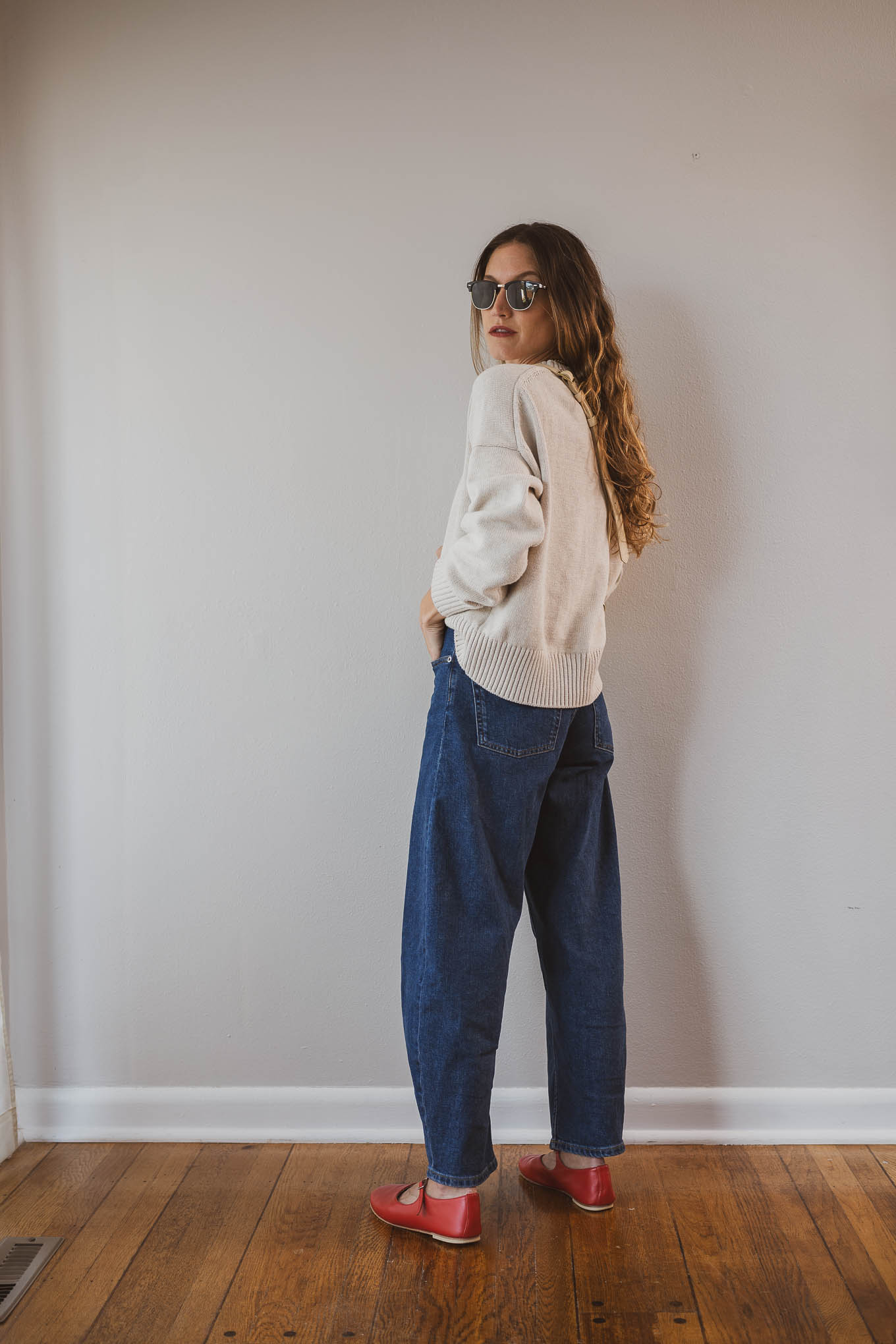 fall capsule wardrobe outfit with a beige cotton sweater, barrel jeans, red mary jane ballet flats, and a cream sling bag