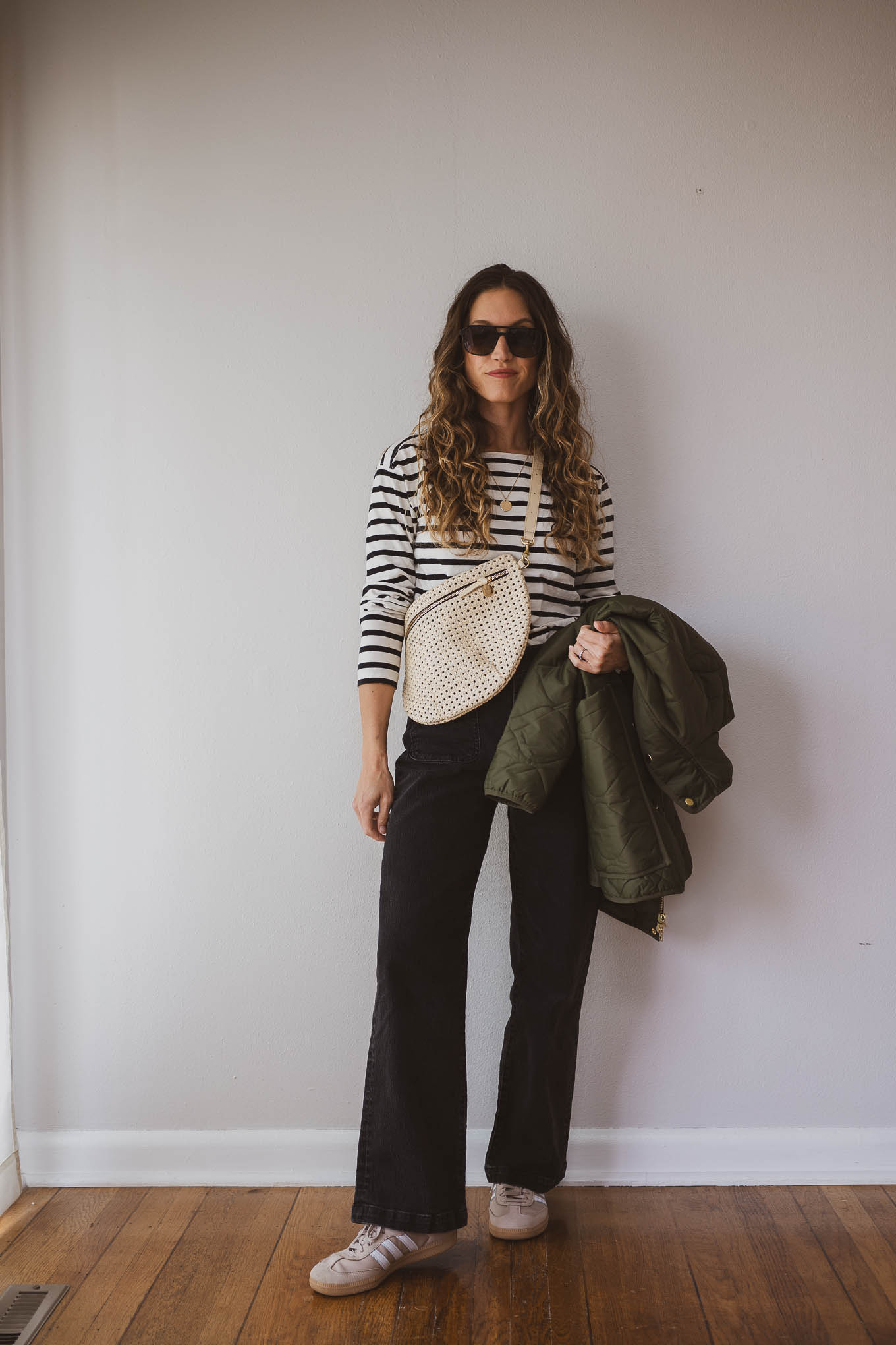 fall outfit idea with a black and white striped top, black straight leg jeans, tan Adidas Samba sneakers, a green quilted jacket and a cream sling bag