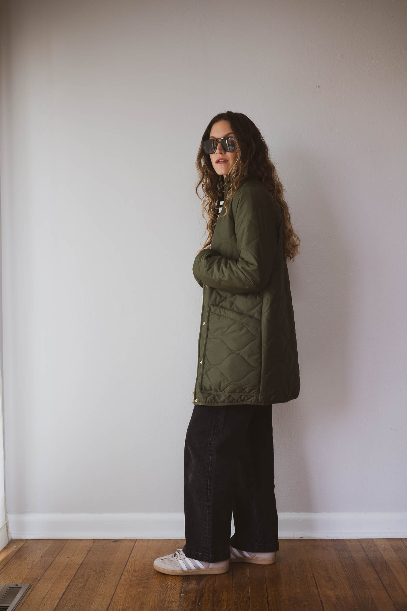 fall outfit idea with a black and white striped top, black straight leg jeans, tan Adidas Samba sneakers, a green quilted jacket and a cream sling bag