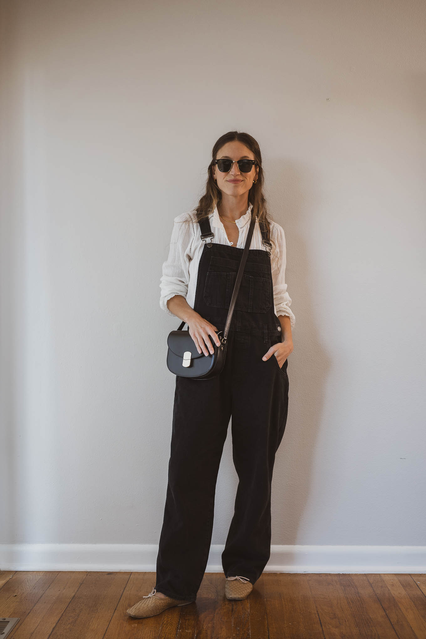 fall capsule wardrobe outfit with a white blouse, black baggy overalls, tan woven ballet flats, and a black crossbody bag
