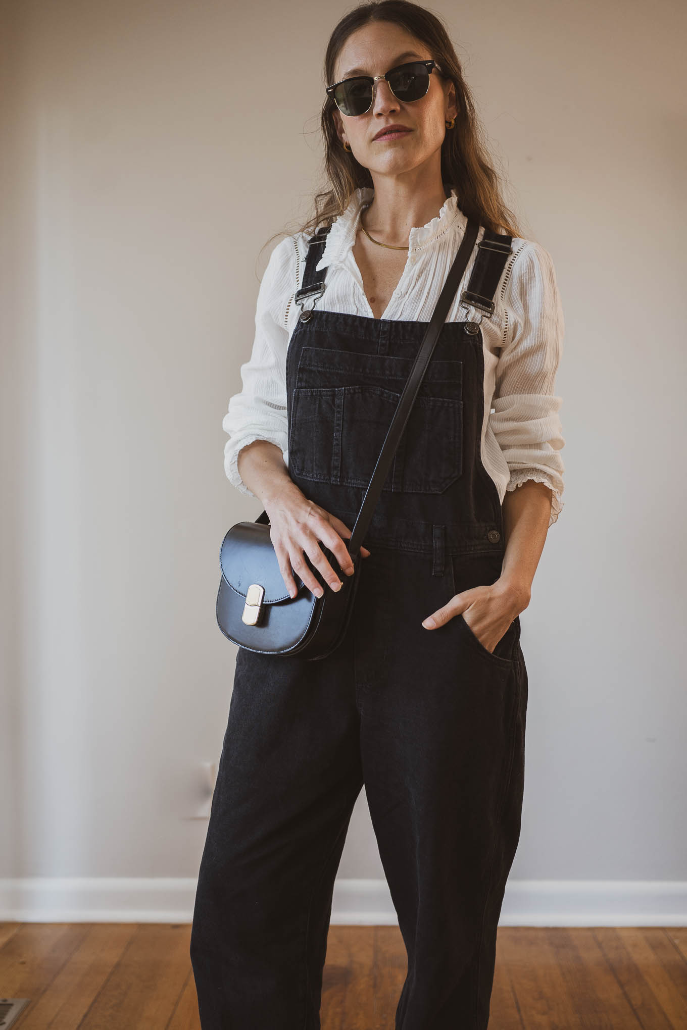 fall capsule wardrobe outfit with a white blouse, black baggy overalls, tan woven ballet flats, and a black crossbody bag