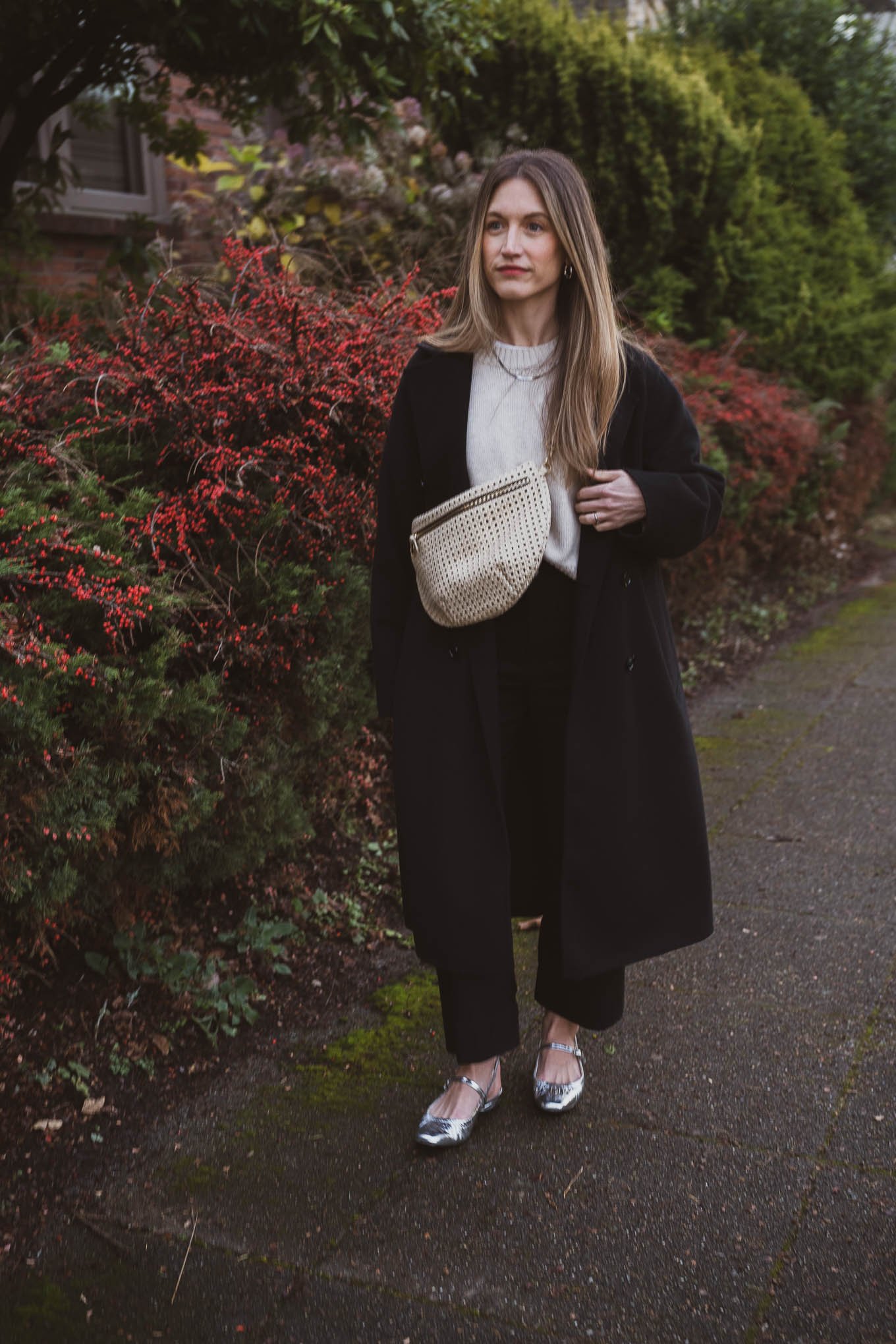 casual chic winter outfit with a beige sweater, black wool coat, black cropped pants, silver ballet flats, and a perforated cream sling bag